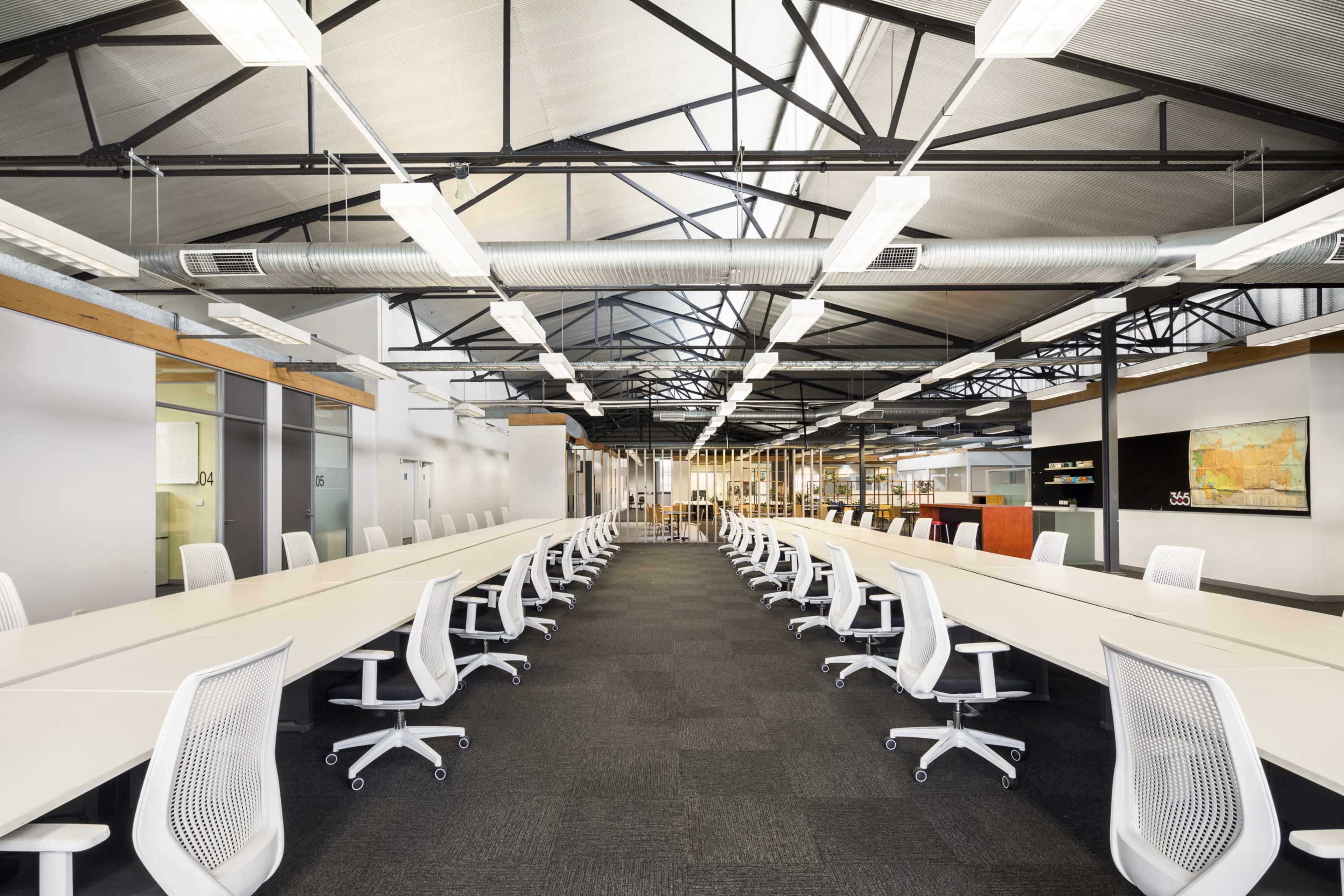 The image shows a modern open-office workspace with long tables and white ergonomic chairs arranged in a symmetrical layout under a high, vaulted ceiling.