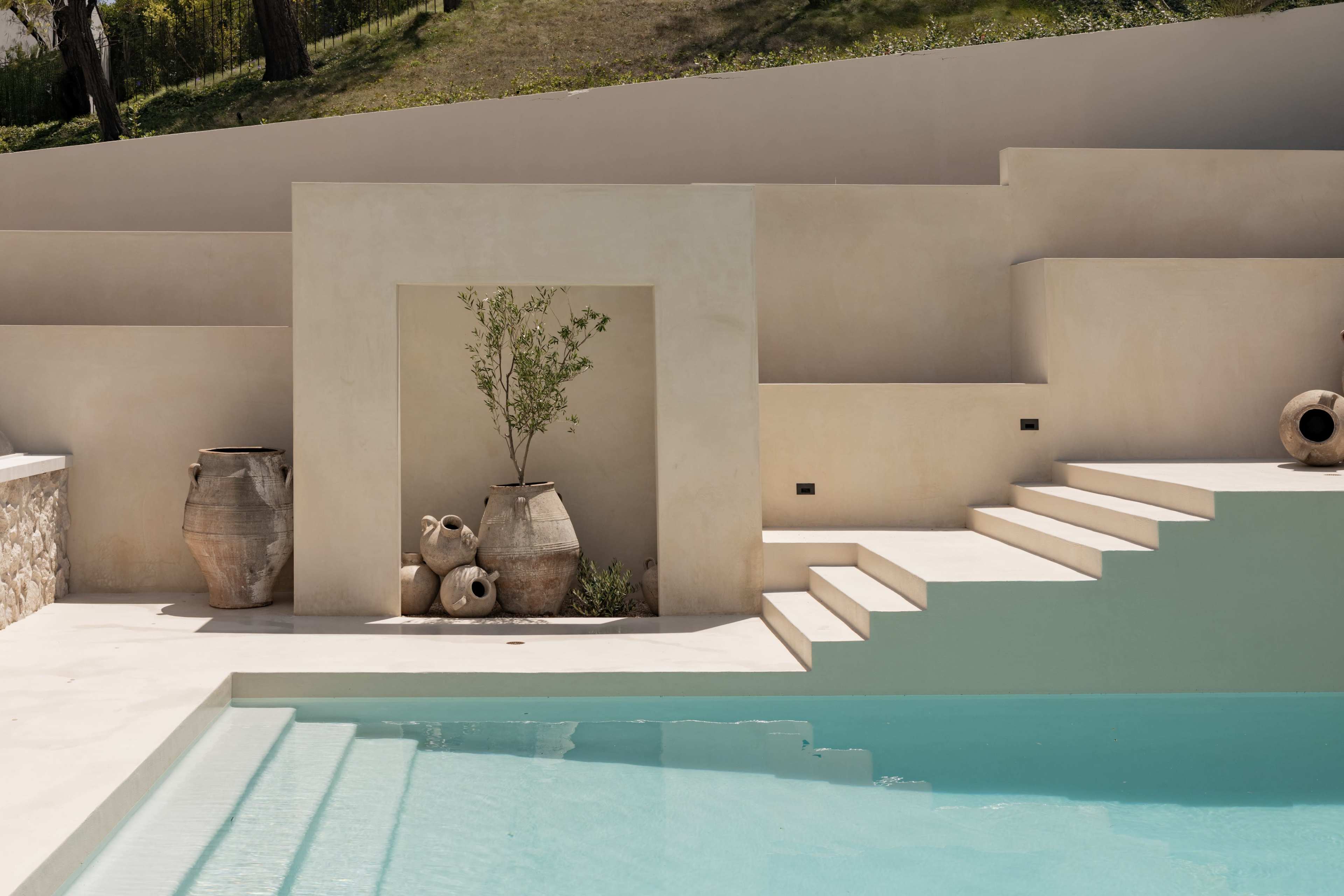 A minimalistic pool area features textured walls, stone steps, and potted plants arranged beside large clay pots.