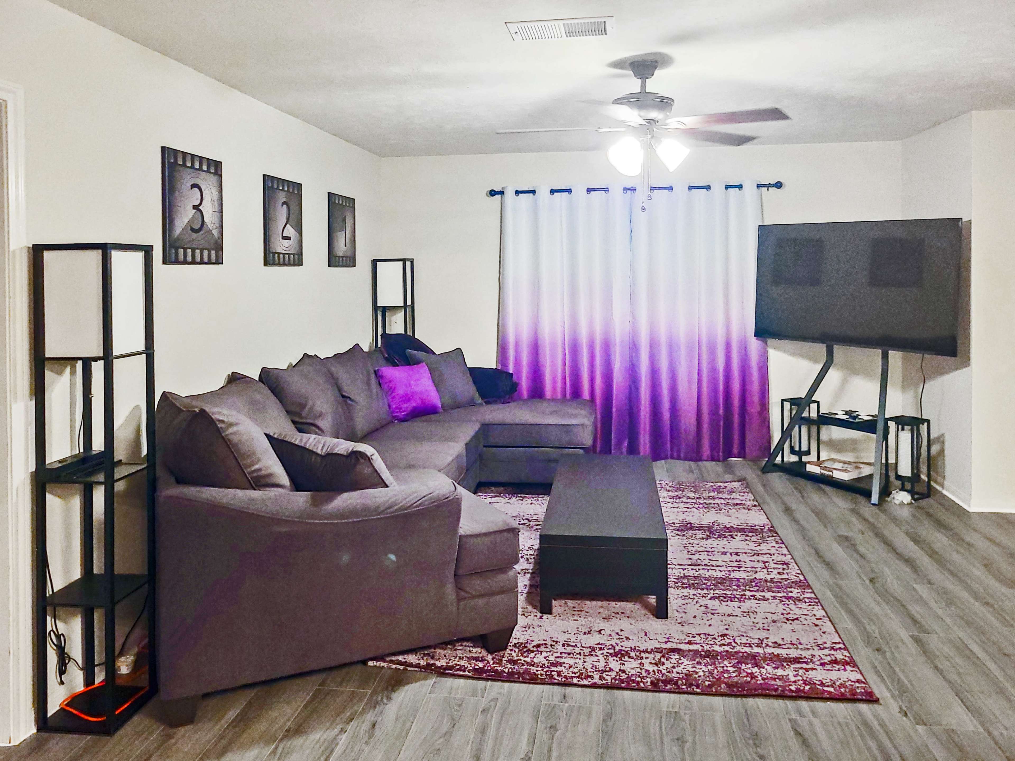 A modern living room features a large gray sectional sofa, a dark coffee table, an area rug, and a television on a stand, complemented by curtains that gradient from white to purple.