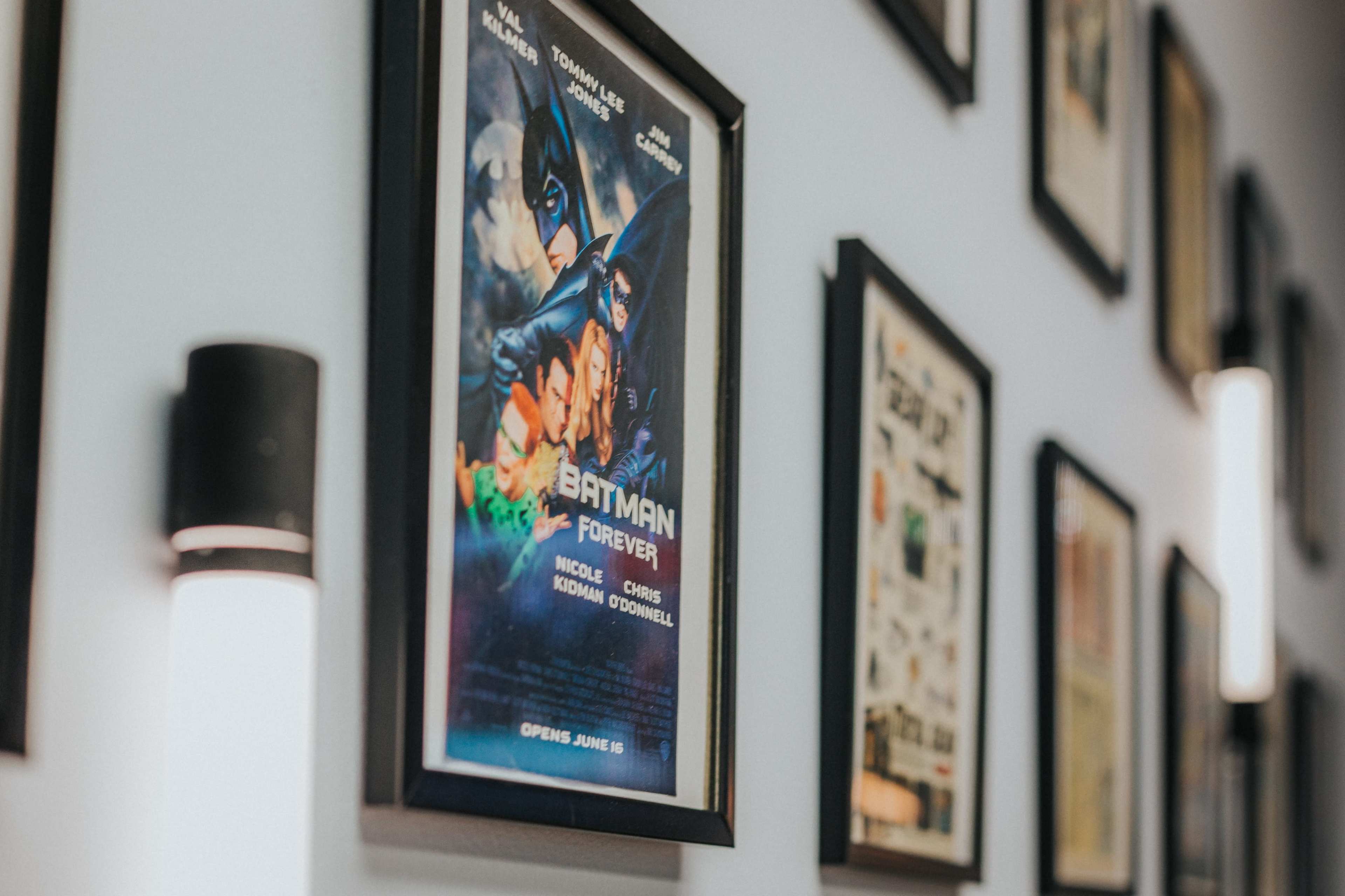 A framed movie poster for "Batman Forever" hangs on a wall adorned with various other framed artworks and articles.