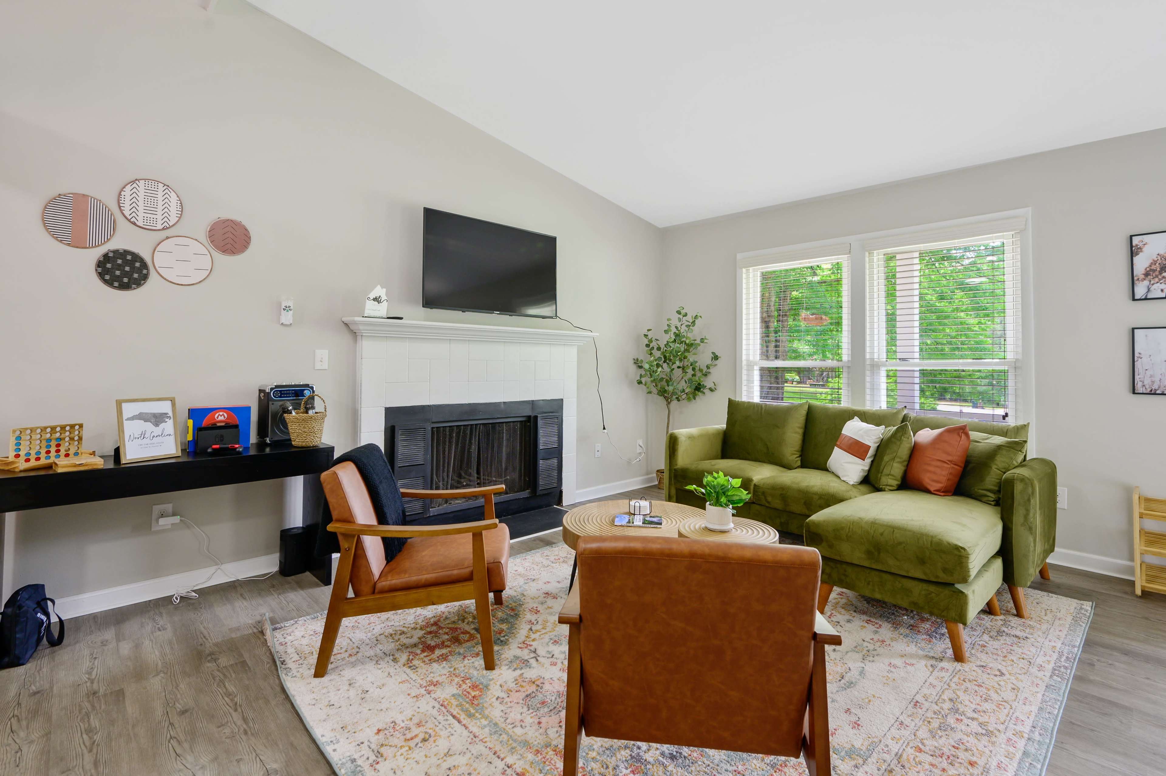 The image shows a modern living room featuring a green sectional sofa, a TV mounted above a white fireplace, and two brown armchairs, all arranged around a round coffee table on a patterned rug.