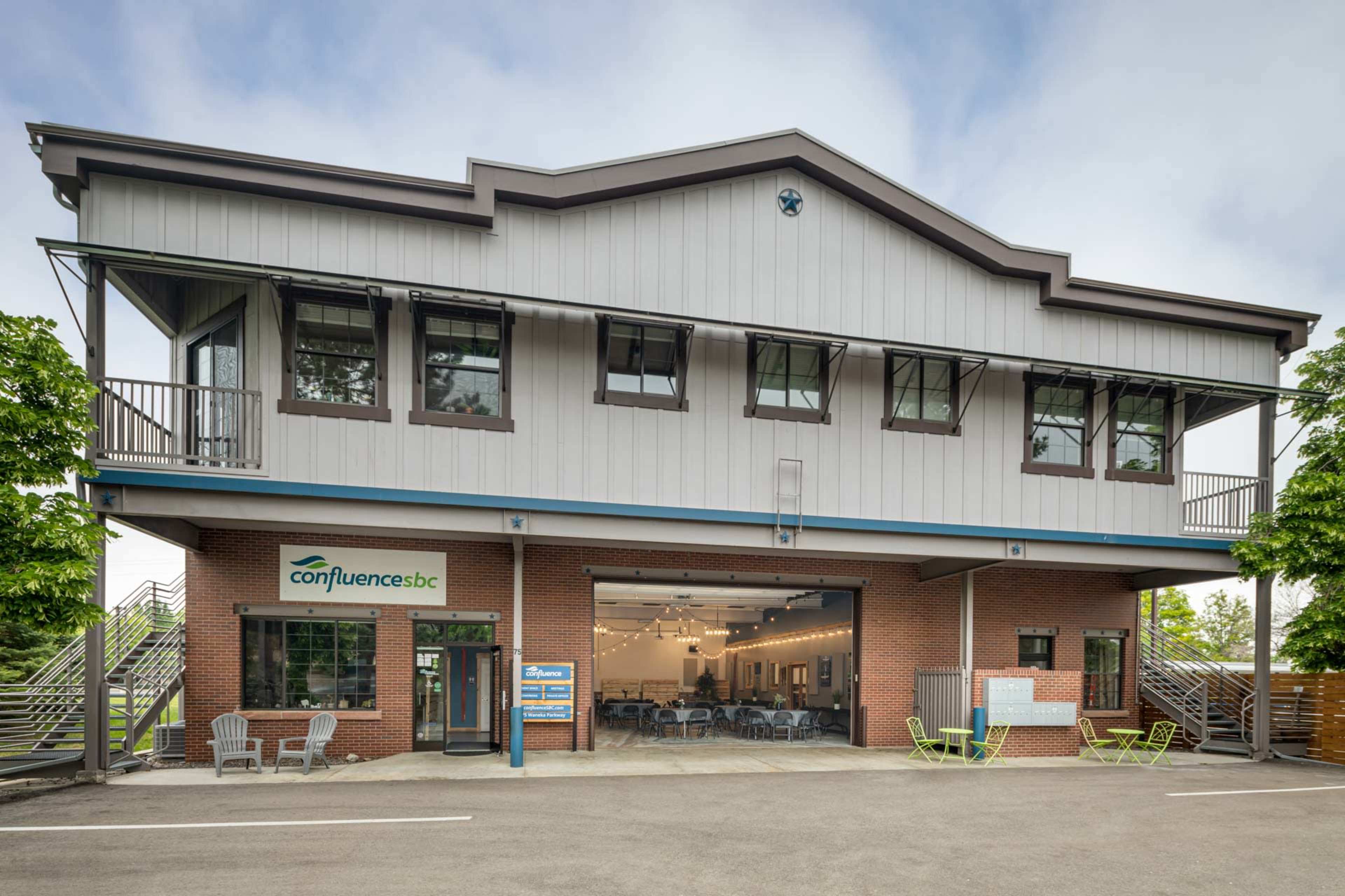 The image shows a two-story building with a modern design, featuring large windows and a porch area with outdoor seating.