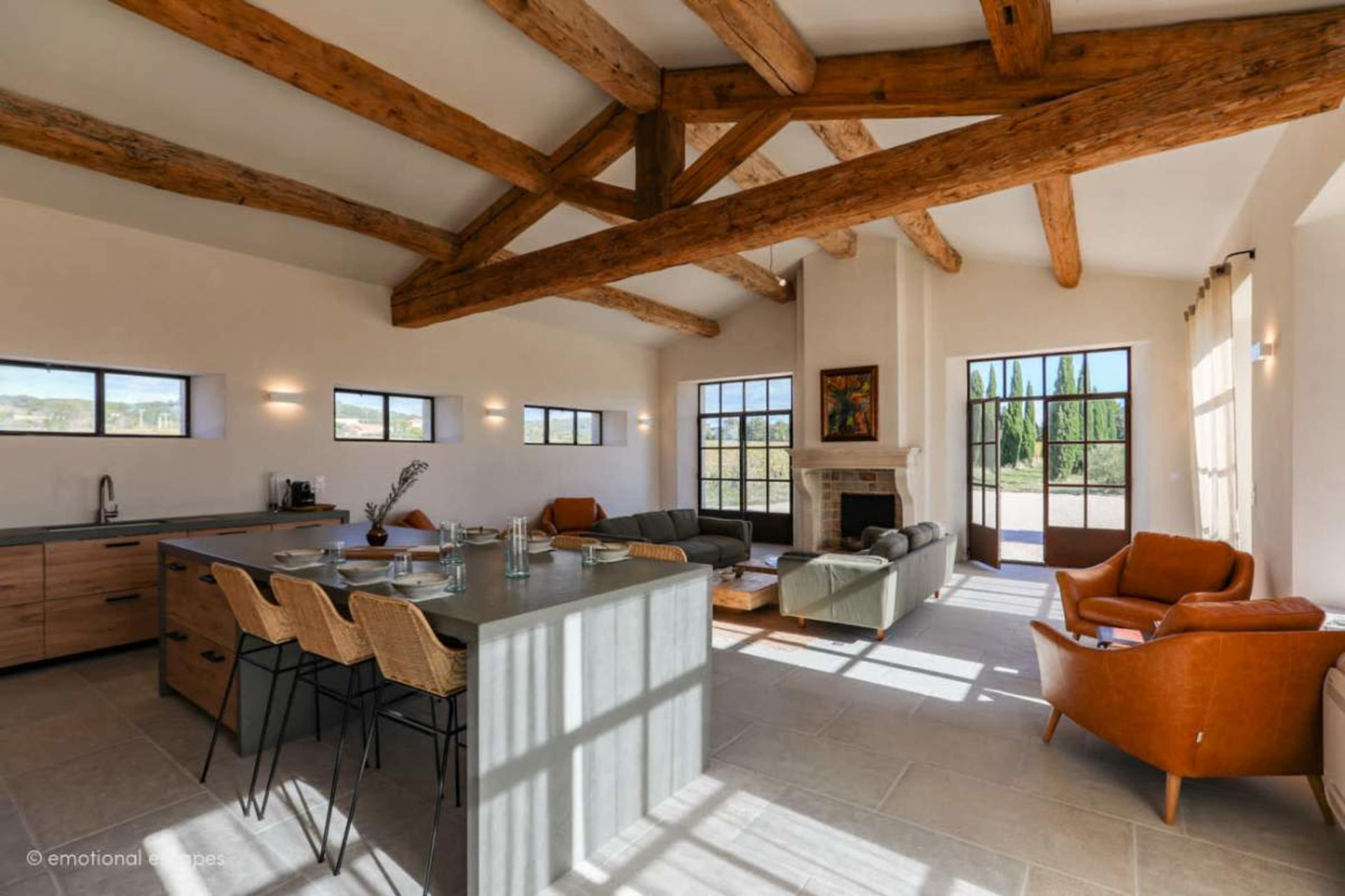 The image shows a spacious living area with high wooden beams, large windows, and modern furniture including a gray sofa, orange chairs, and a stone fireplace.
