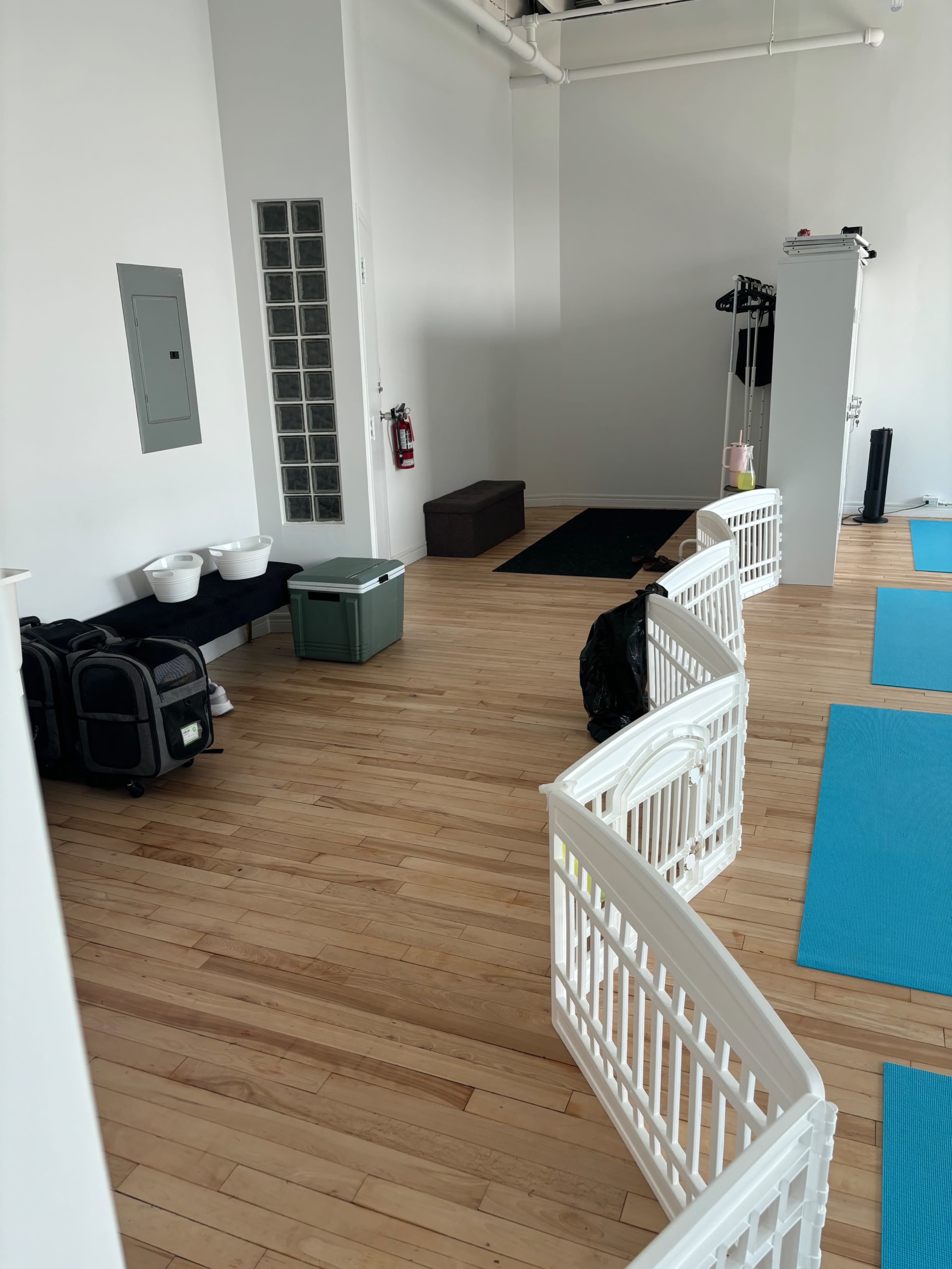 A spacious, well-lit room with wooden flooring, featuring a partition of white safety gates, storage bins, and yoga mats on the floor.