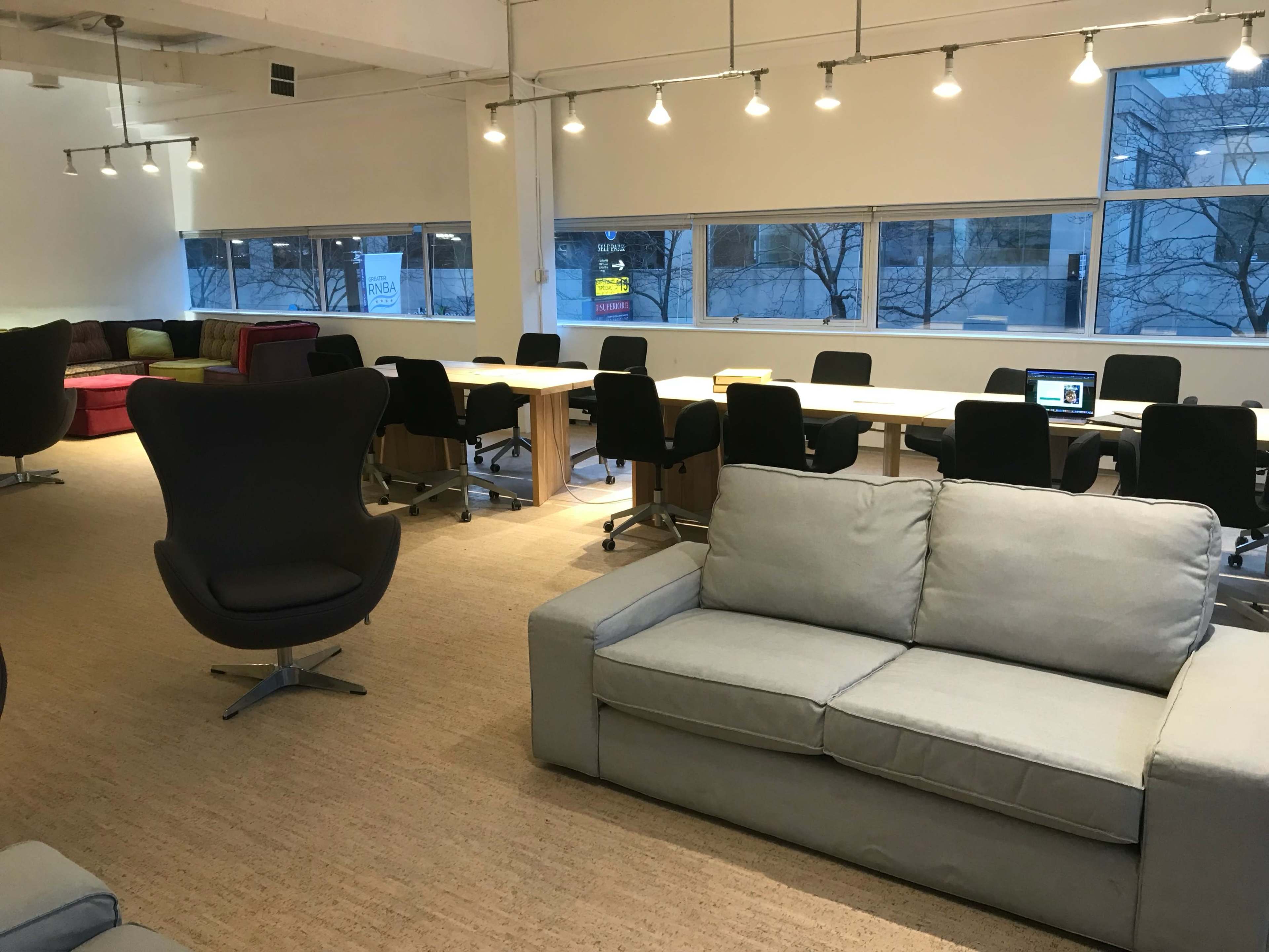 A spacious office area with a mix of seating arrangements, including a gray sofa, black chairs, and long tables beneath overhead lighting.