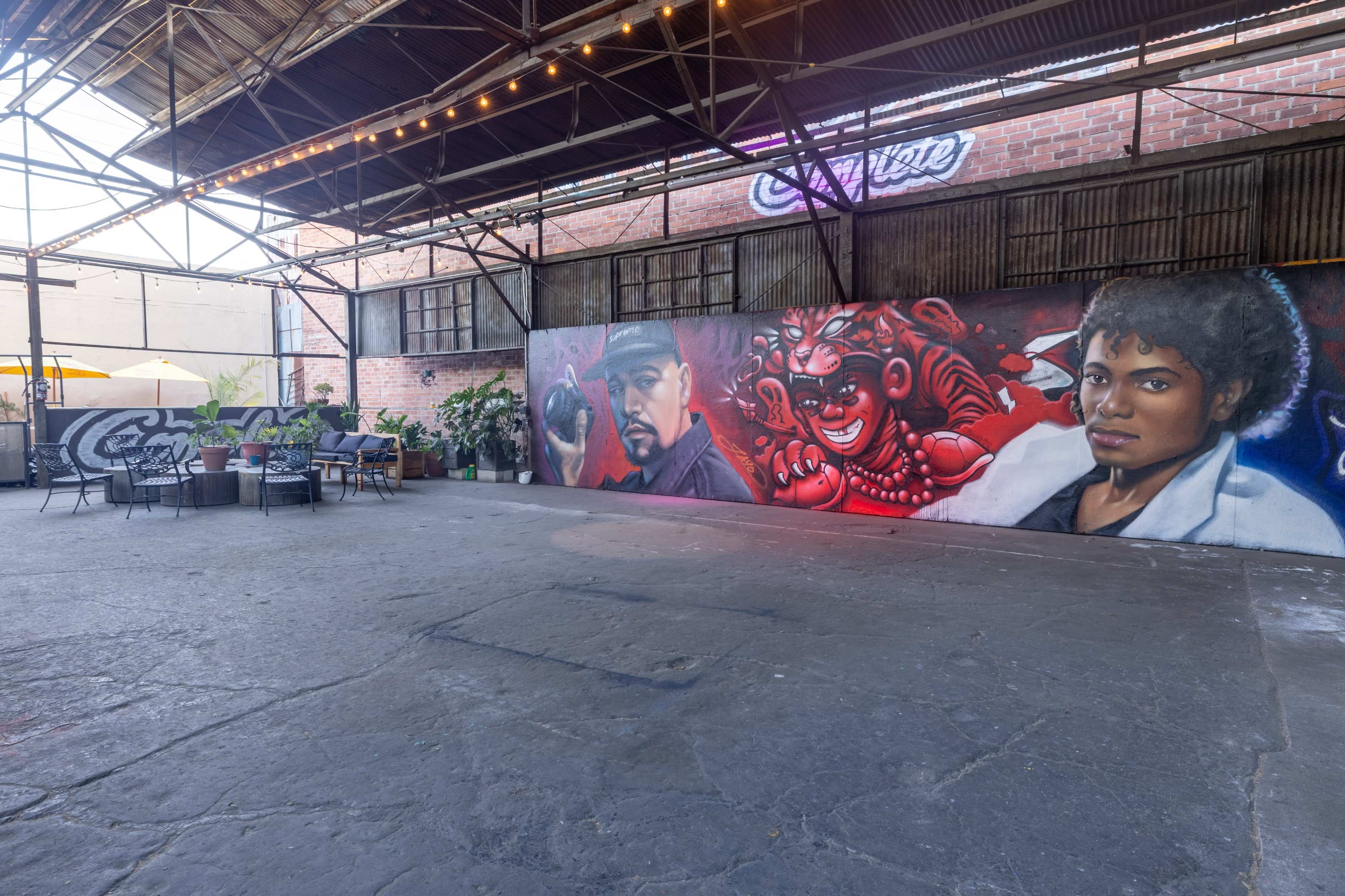 The image shows an industrial-style interior space featuring a large mural of three figures on a brick wall, with seating arrangements and potted plants nearby under overhead string lights.