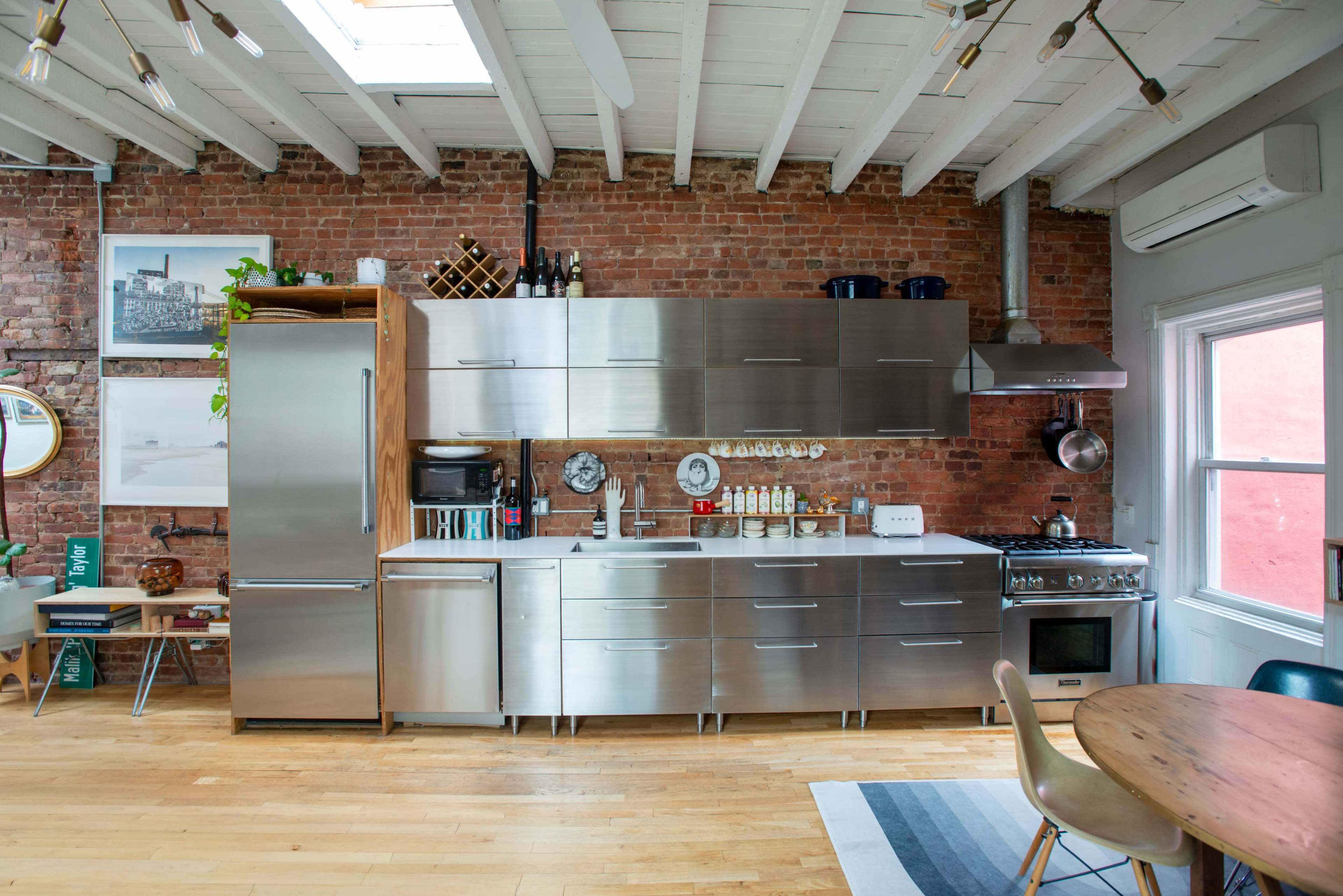 The image shows a modern kitchen with stainless steel cabinets, open shelving, a brick wall, and wooden flooring.