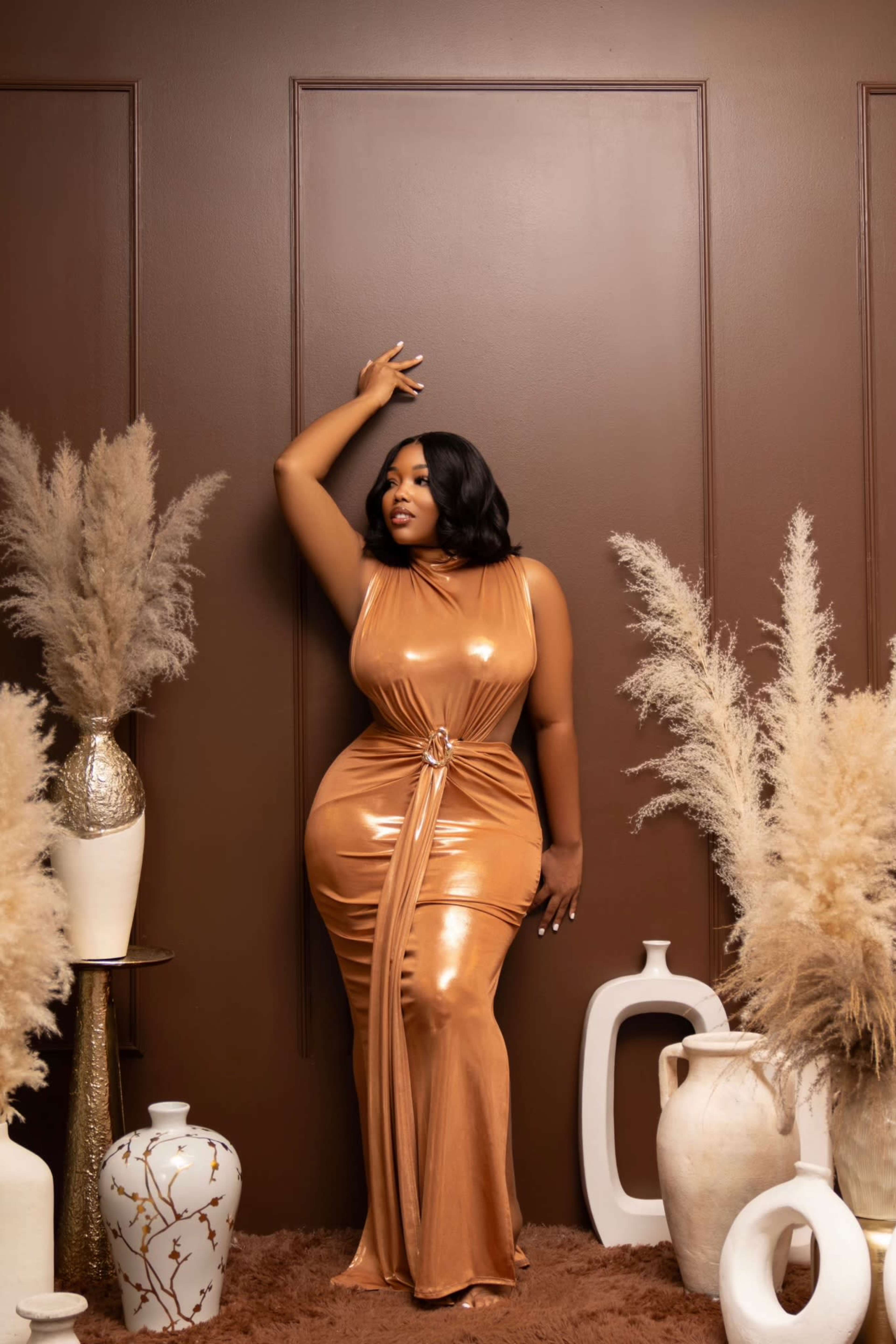 A woman in a metallic bronze dress poses against a brown wall, surrounded by decorative vases and dried pampas grass.