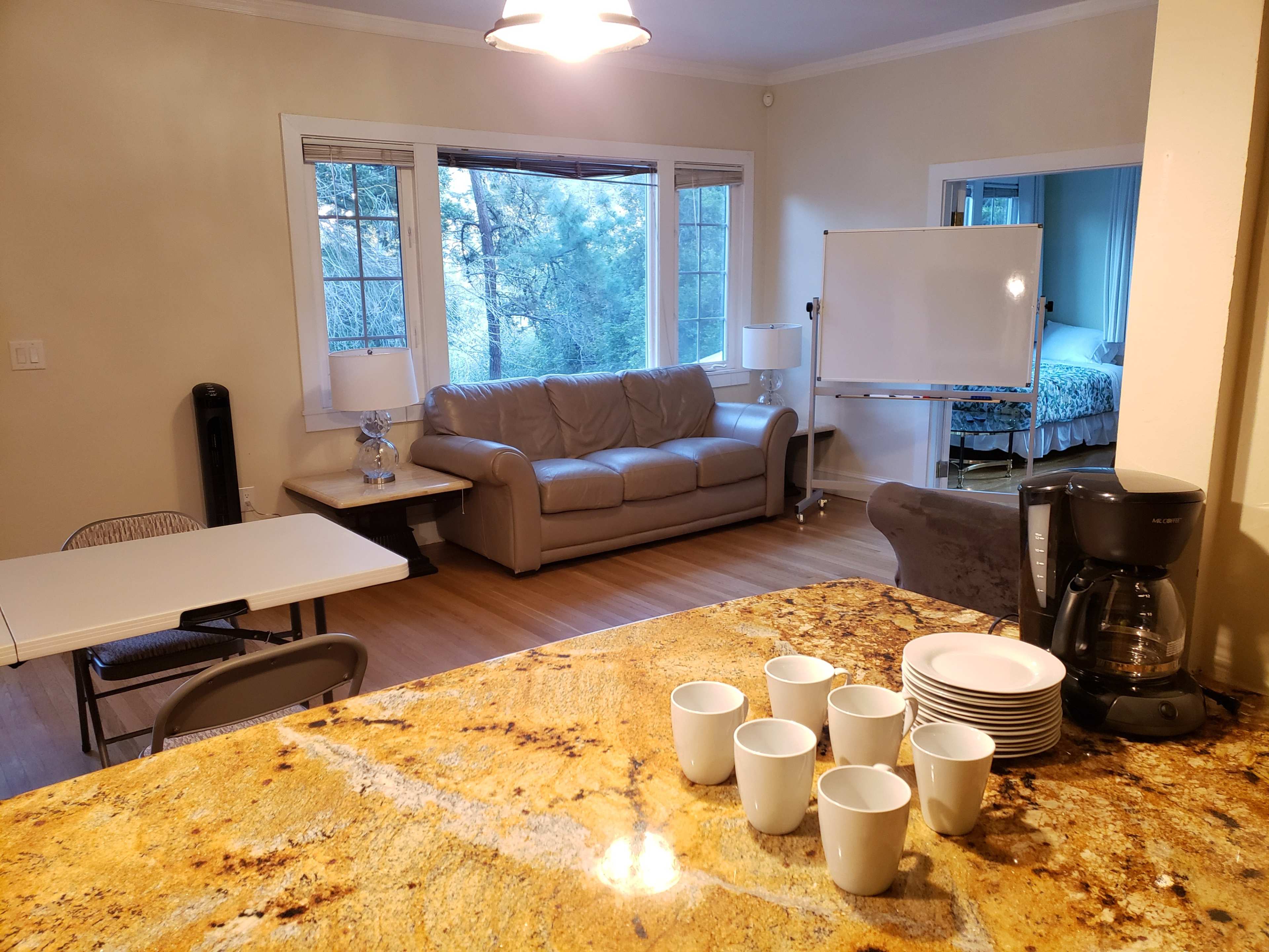 A living room and kitchen area are visible, featuring a couch, a table with chairs, and a coffee maker with several cups arranged on the counter.