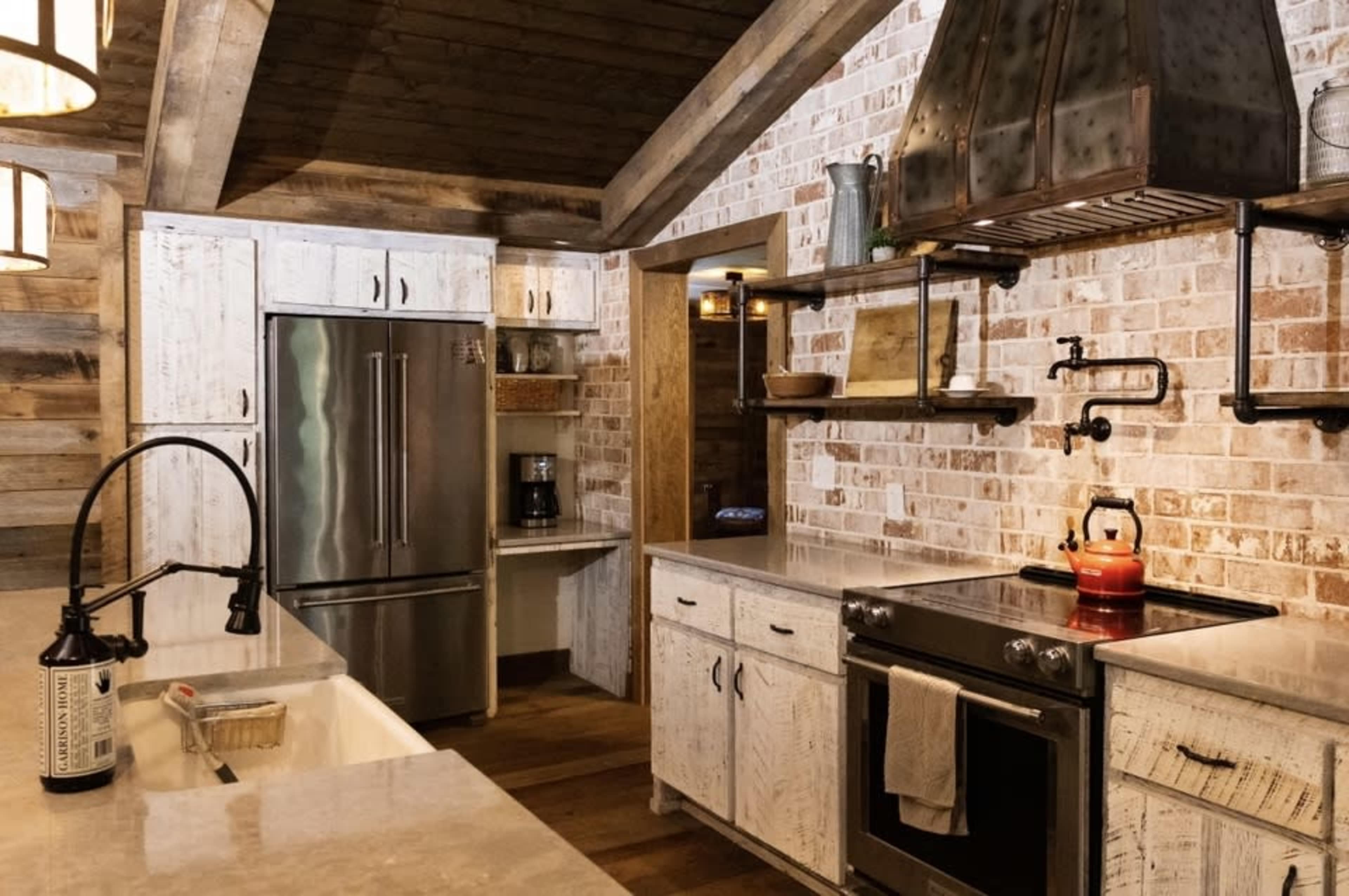 A modern kitchen with white wooden cabinetry, a stainless steel refrigerator, and a gas stove under a metal hood, featuring exposed brick walls and wooden beams.