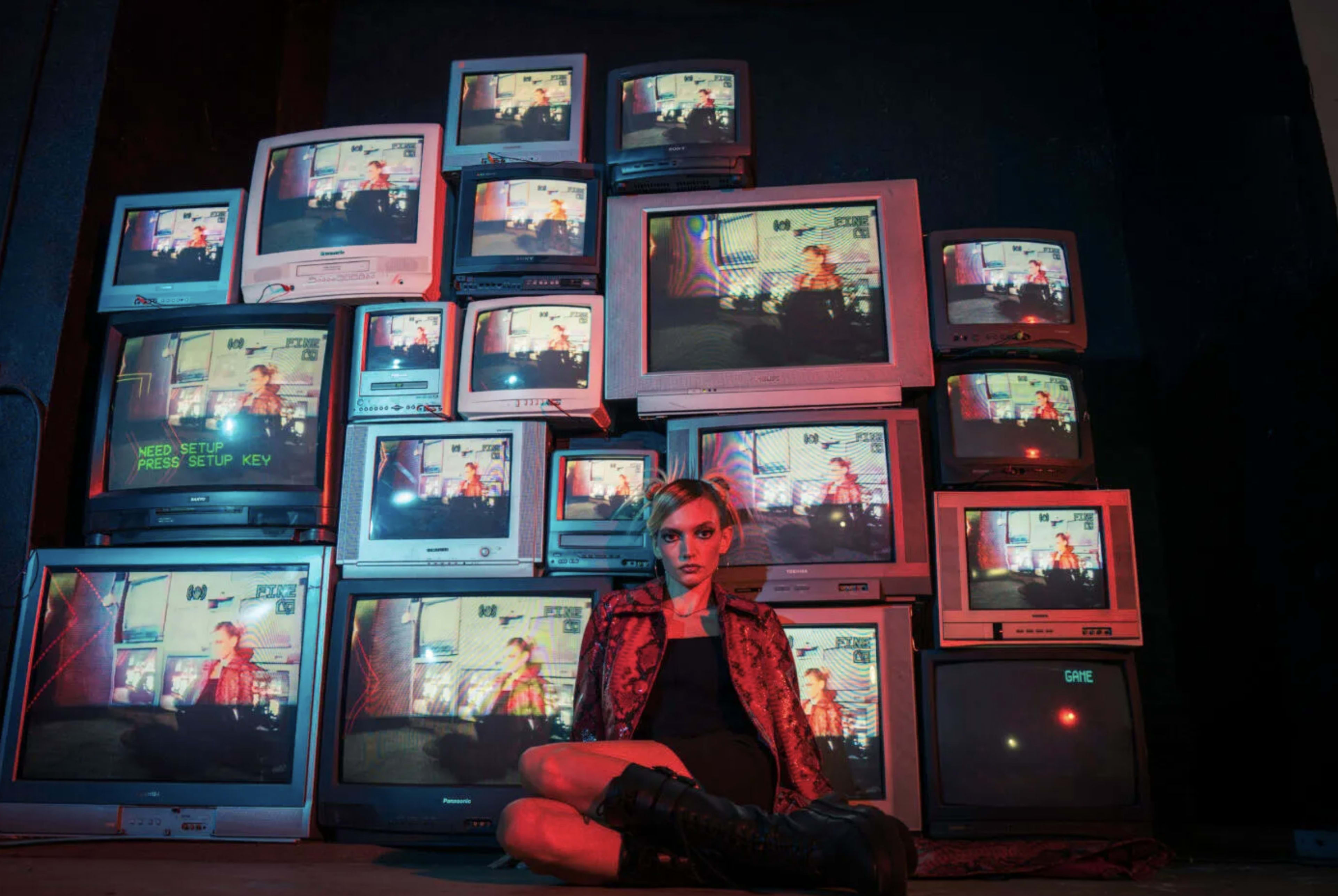 A person sits on the floor in front of a wall of vintage televisions displaying colorful screens.