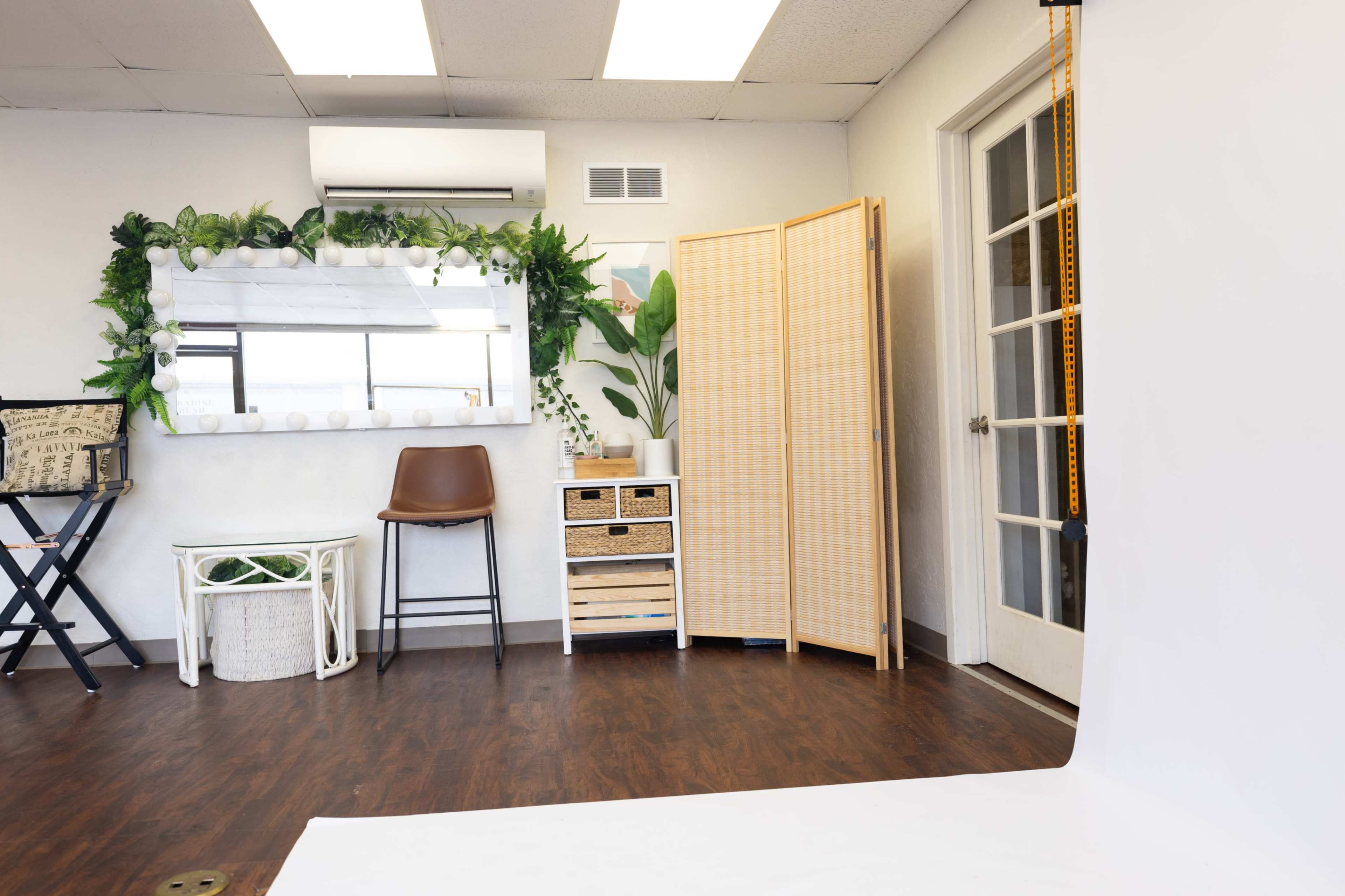 The image shows a well-lit studio space featuring a mirror, a chair, a decorative screen, and plants arranged against a white backdrop.