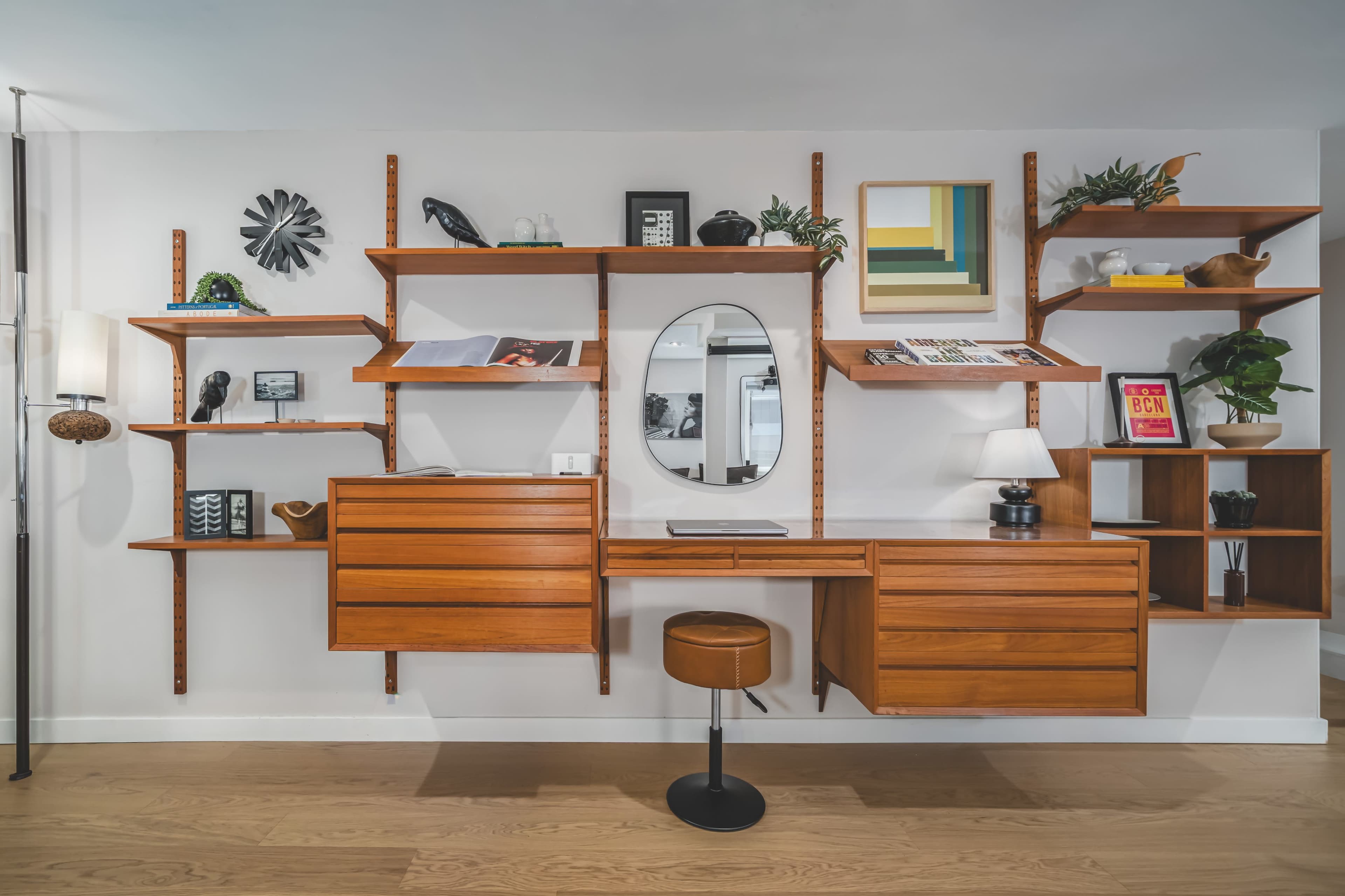 The image shows a modern wooden wall unit featuring shelves, a circular mirror, and a small desk with a stool, all arranged in a bright interior space.