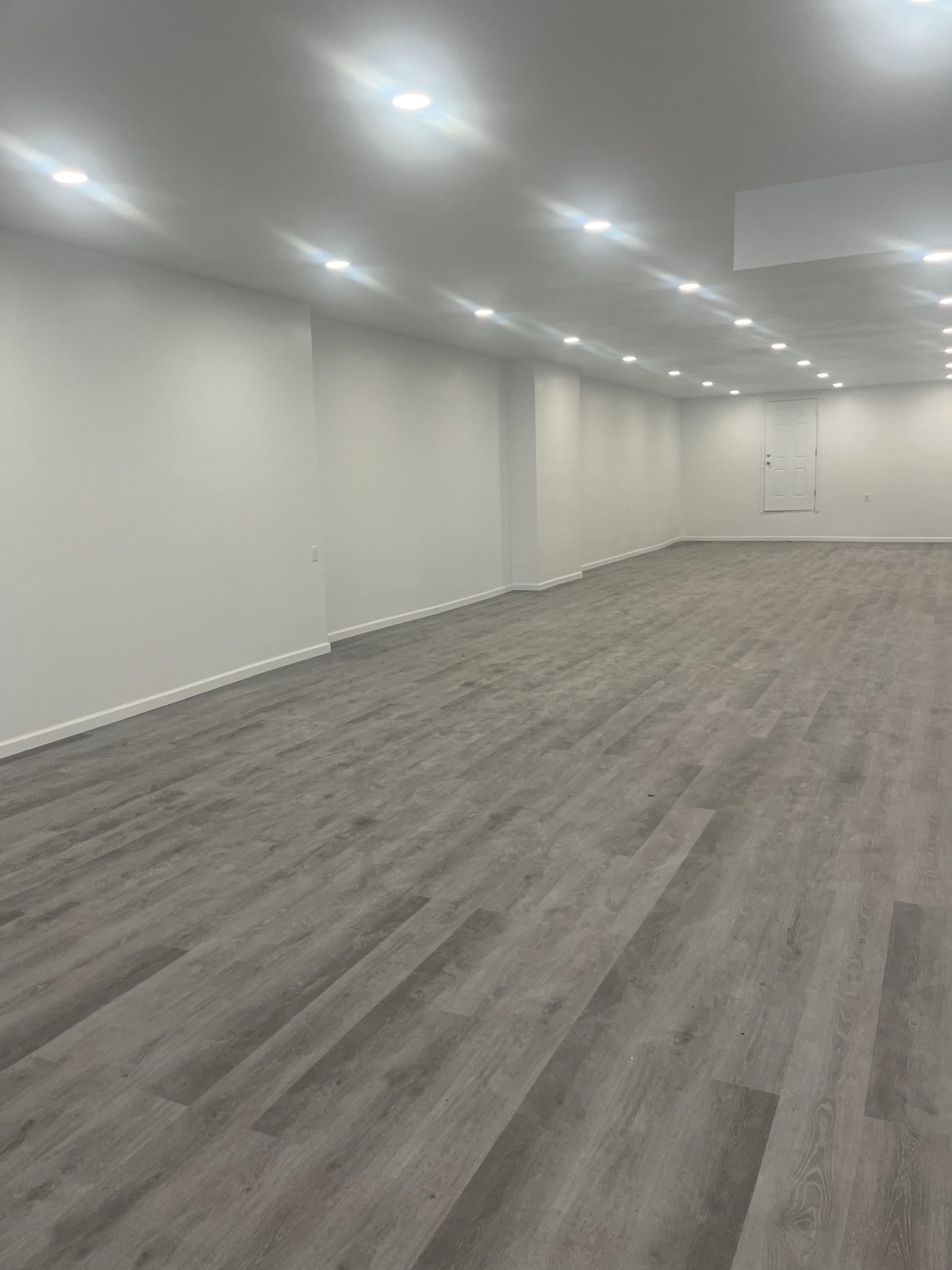 The image shows a large, empty room with gray wood laminate flooring and white walls, illuminated by multiple recessed ceiling lights.