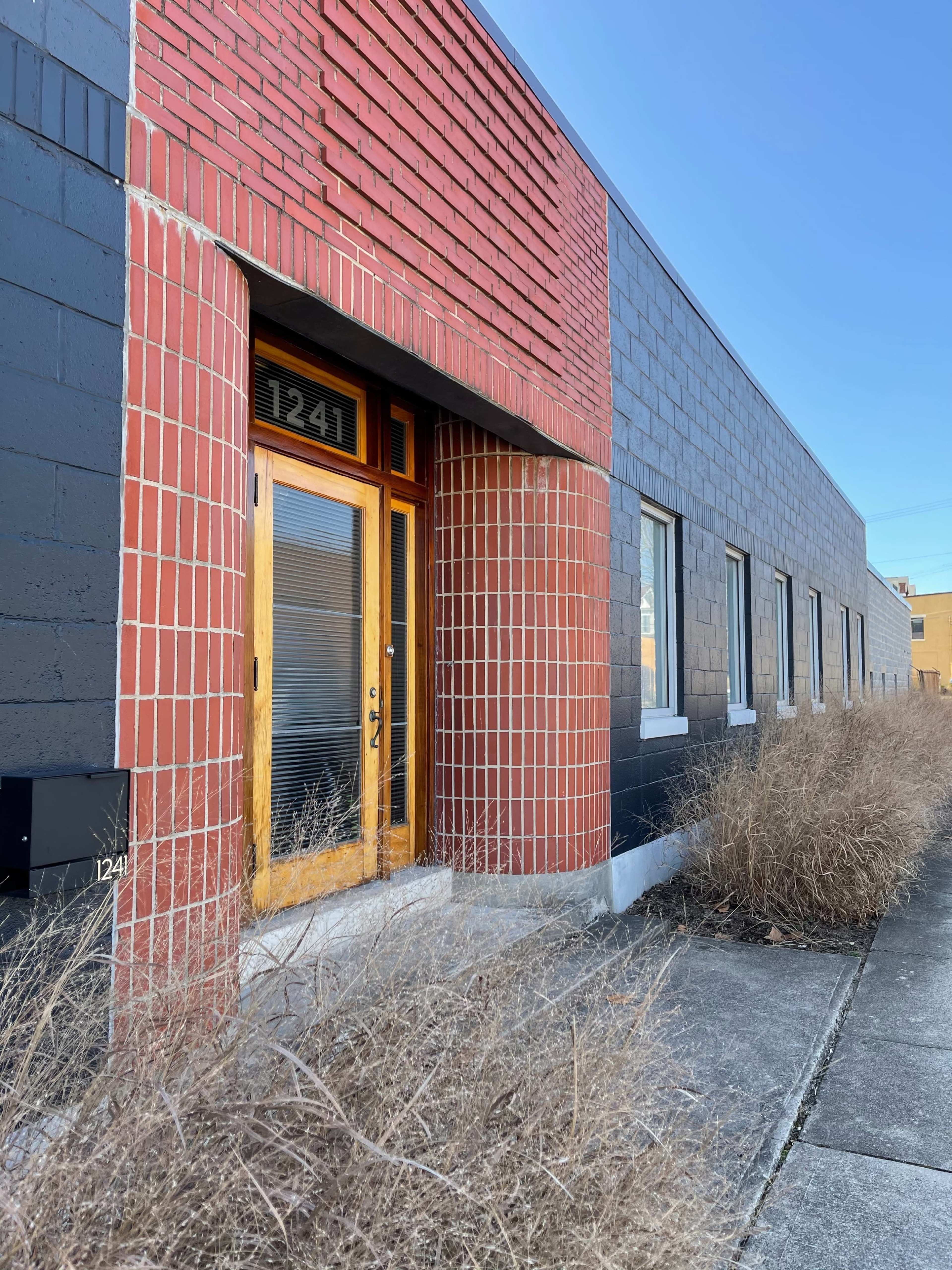 The image shows a building facade featuring a door with a wooden frame, flanked by brick sections in red and gray, amidst overgrown grass along the sidewalk.