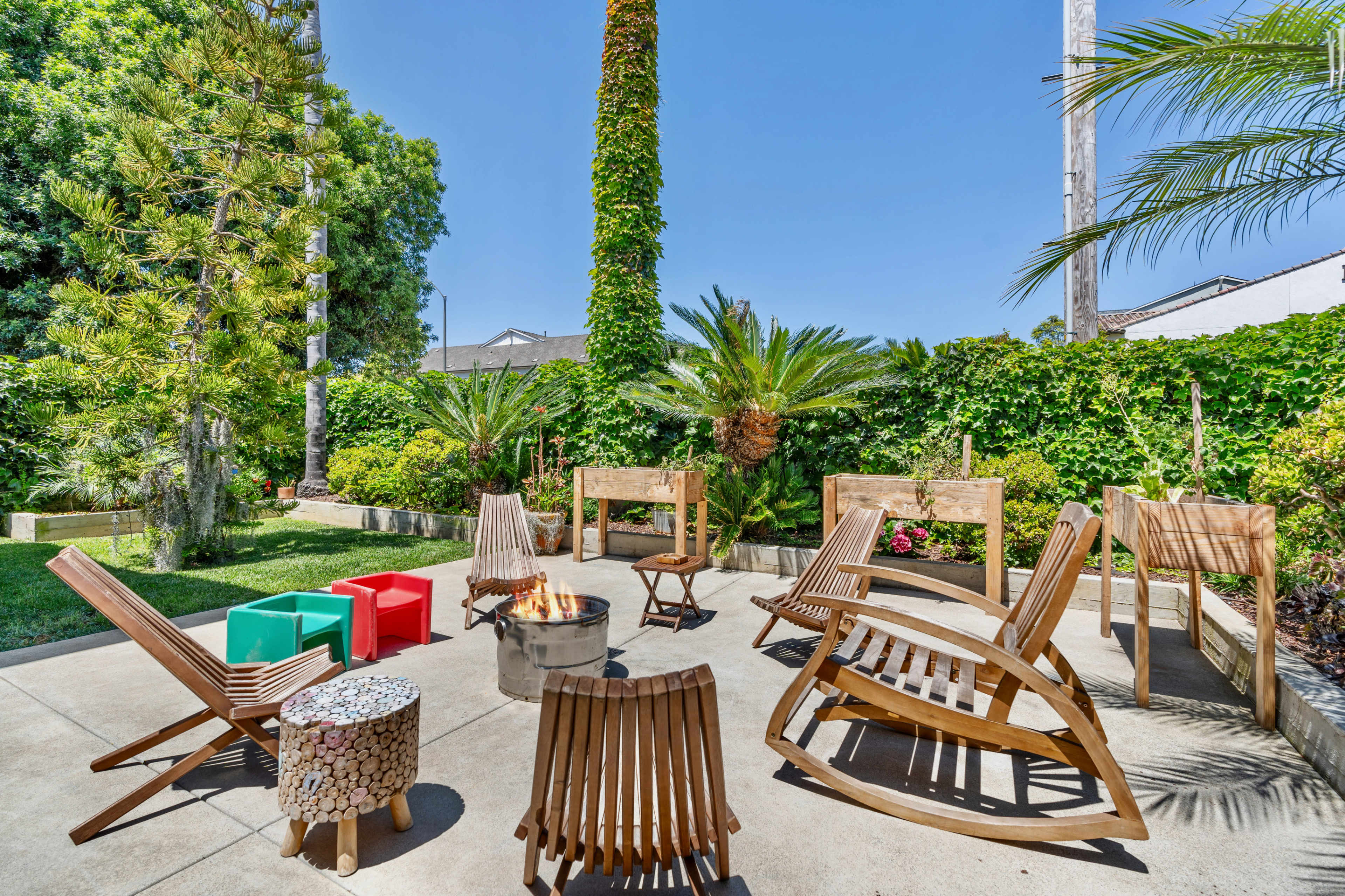 A patio area features wooden chairs and a fire pit surrounded by lush greenery and tall palm trees.