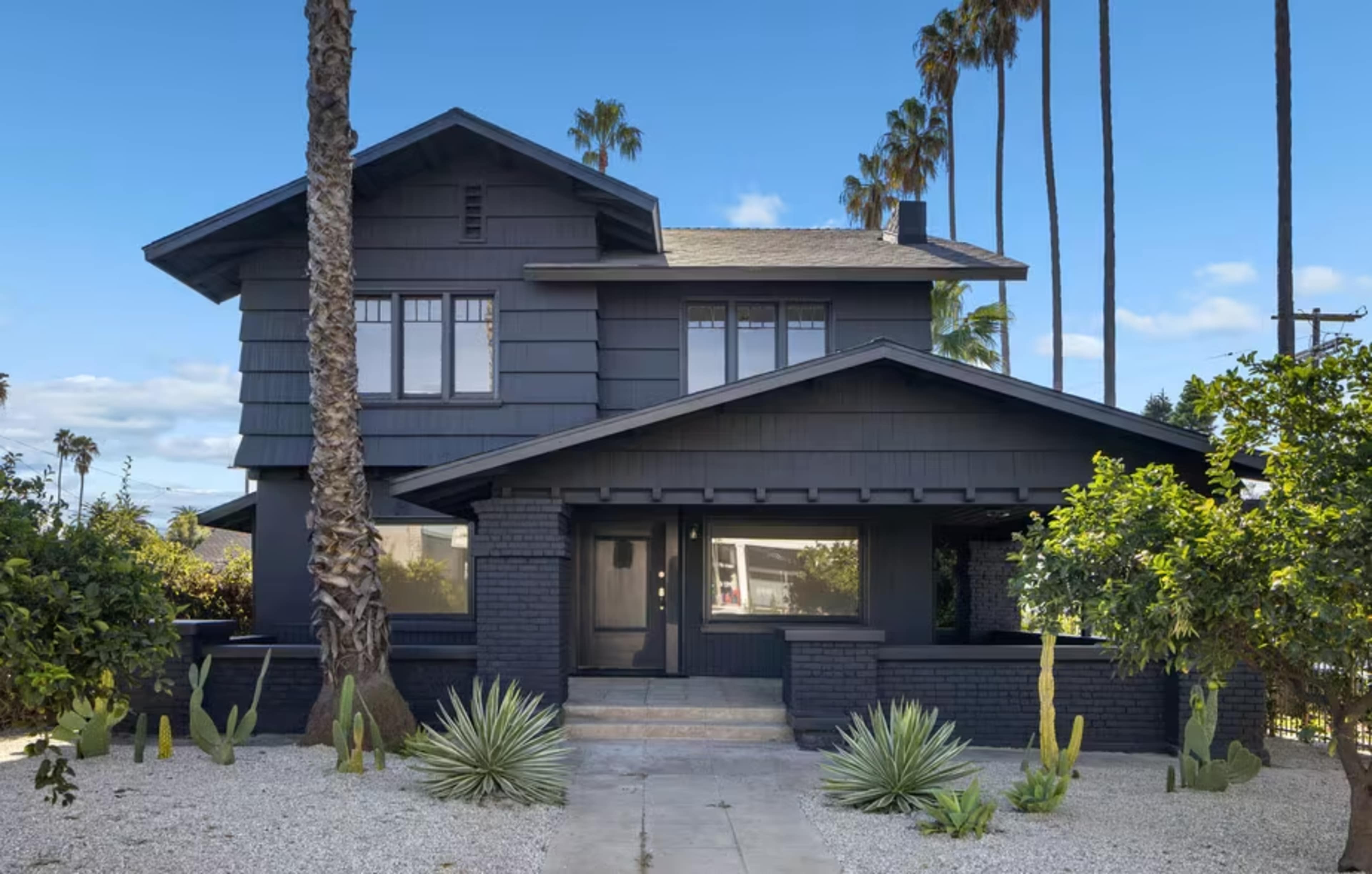The image shows a two-story dark-colored house surrounded by palm trees and desert landscaping, with a pathway leading to the entrance.