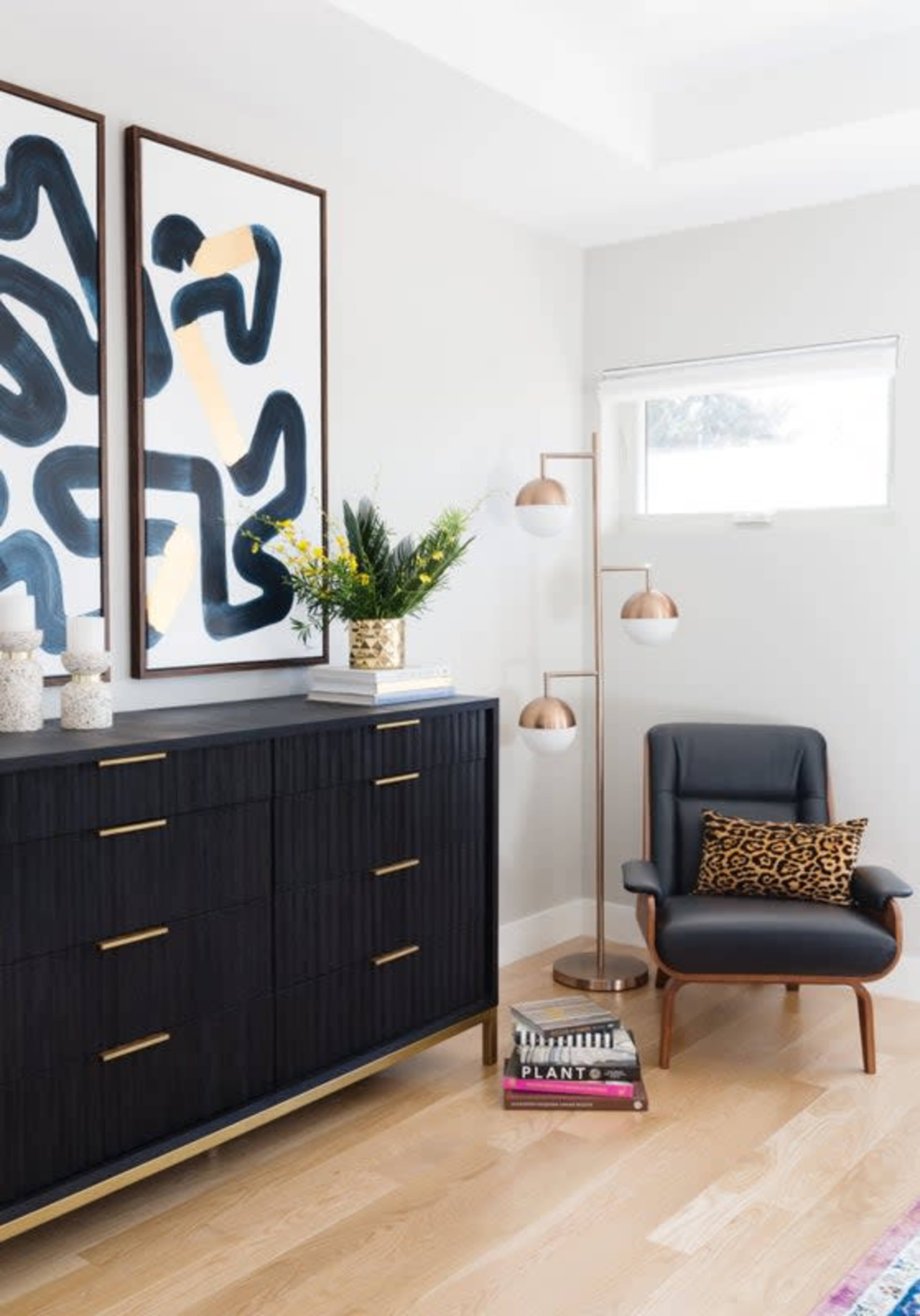 The image shows a stylish corner of a room featuring a dark wooden dresser with gold handles, framed abstract art on the walls, a modern floor lamp, and a comfortable chair with a leopard print cushion.