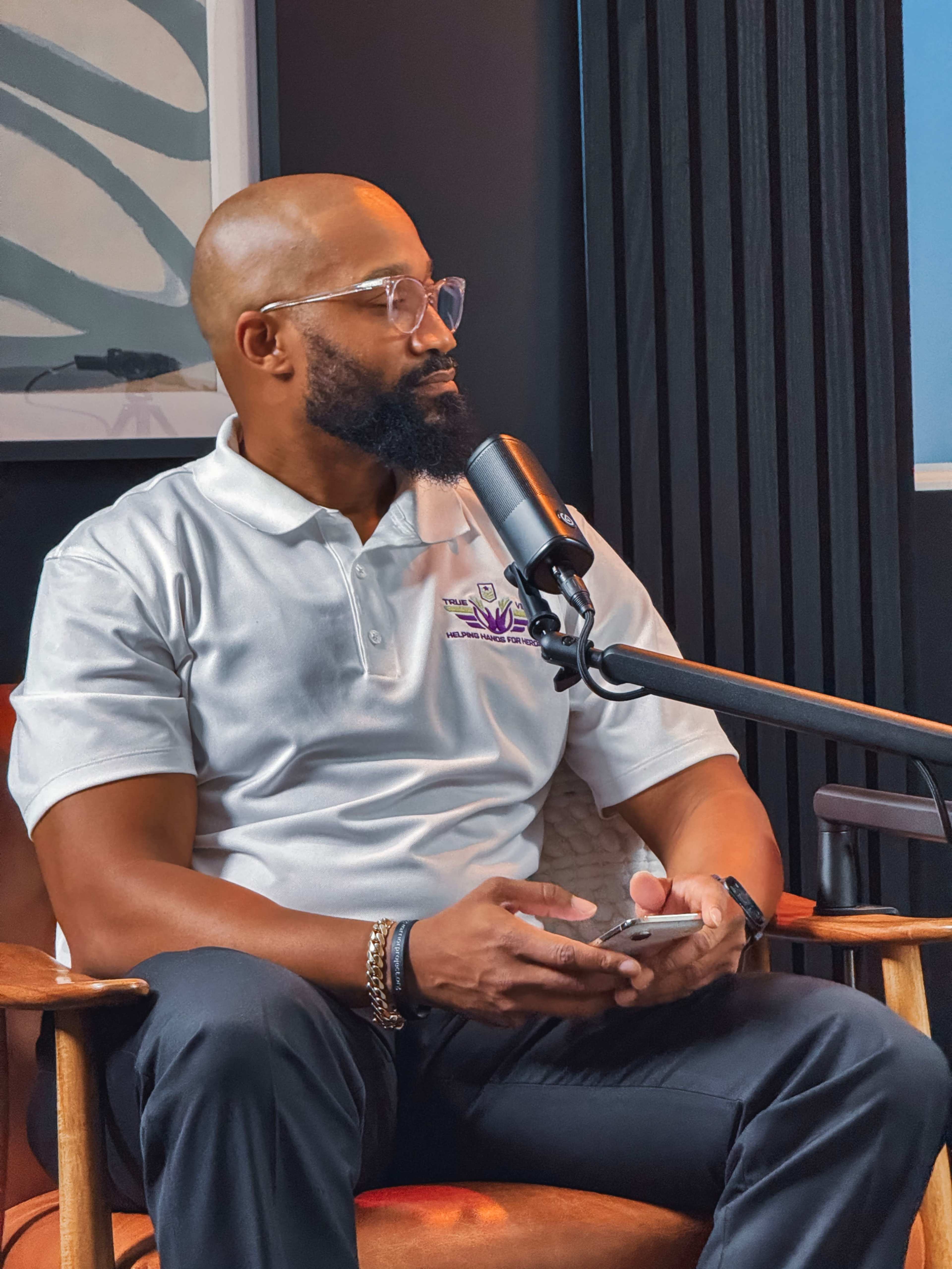 A man wearing glasses and a polo shirt sits in a chair with a microphone in front of him, holding a phone.