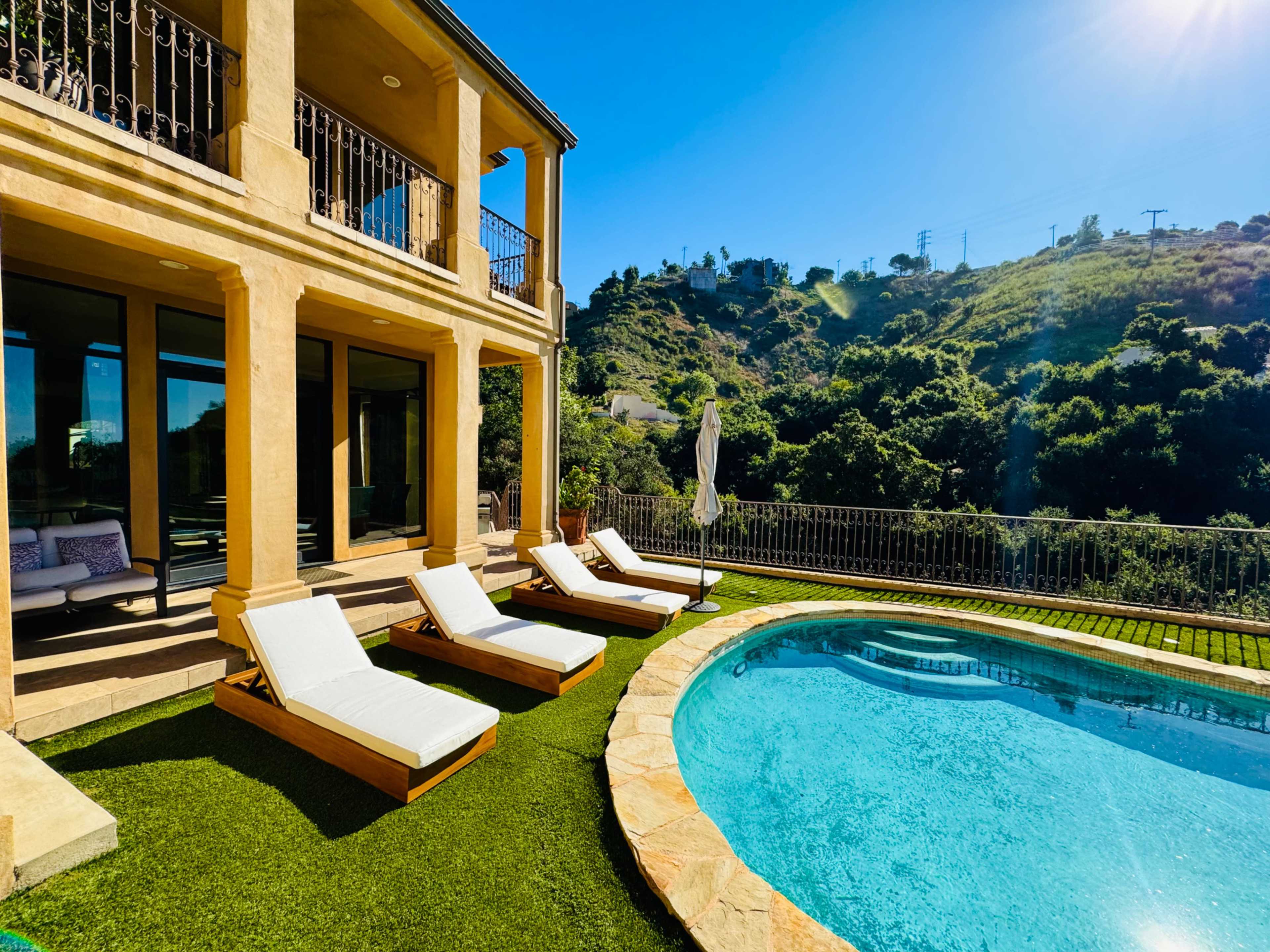 The image shows a modern house with a swimming pool and lounge chairs situated on a grassy area overlooking a hillside.