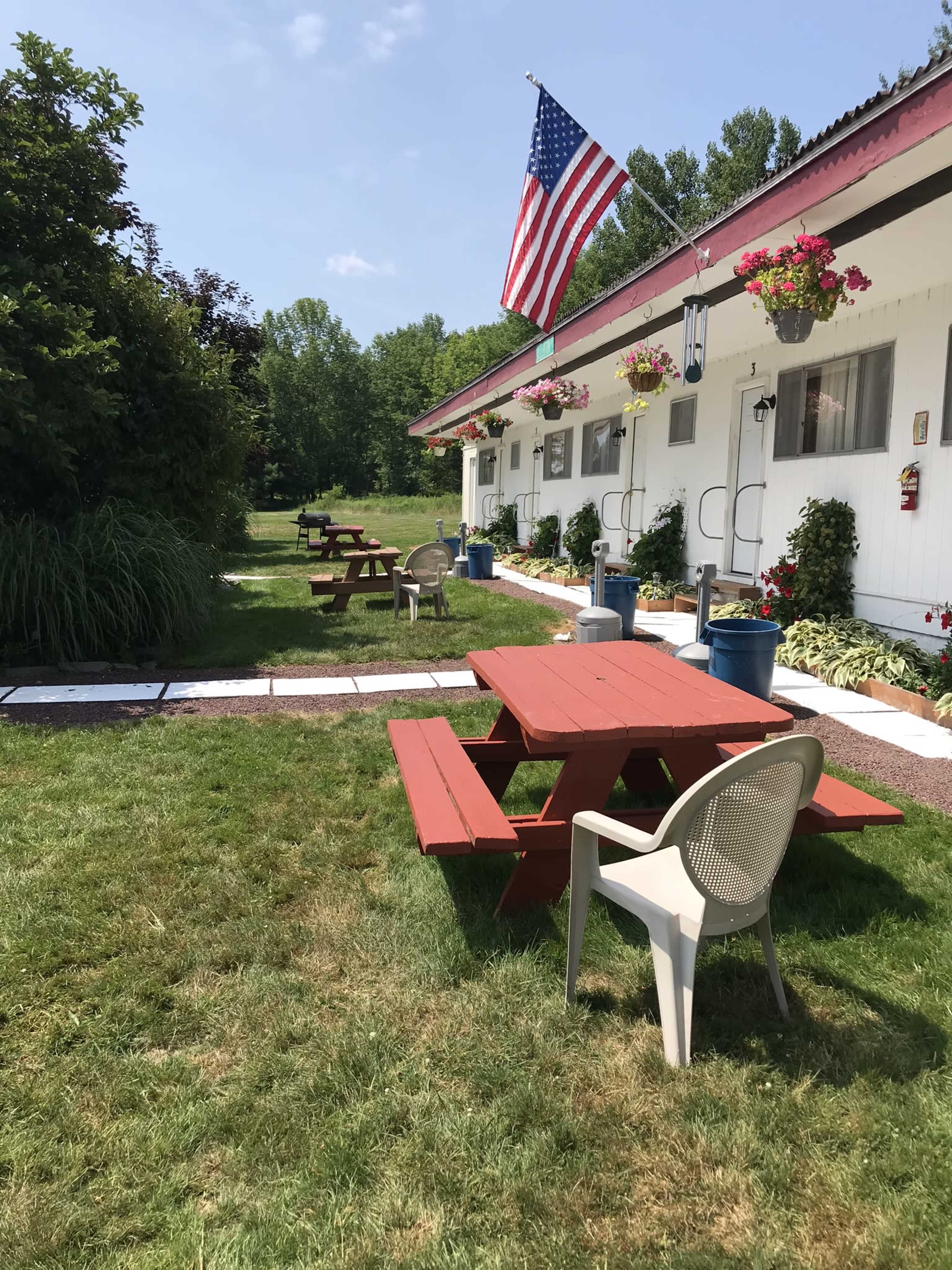 A grassy area with red picnic tables and a white building, featuring an American flag and flower pots along the exterior.