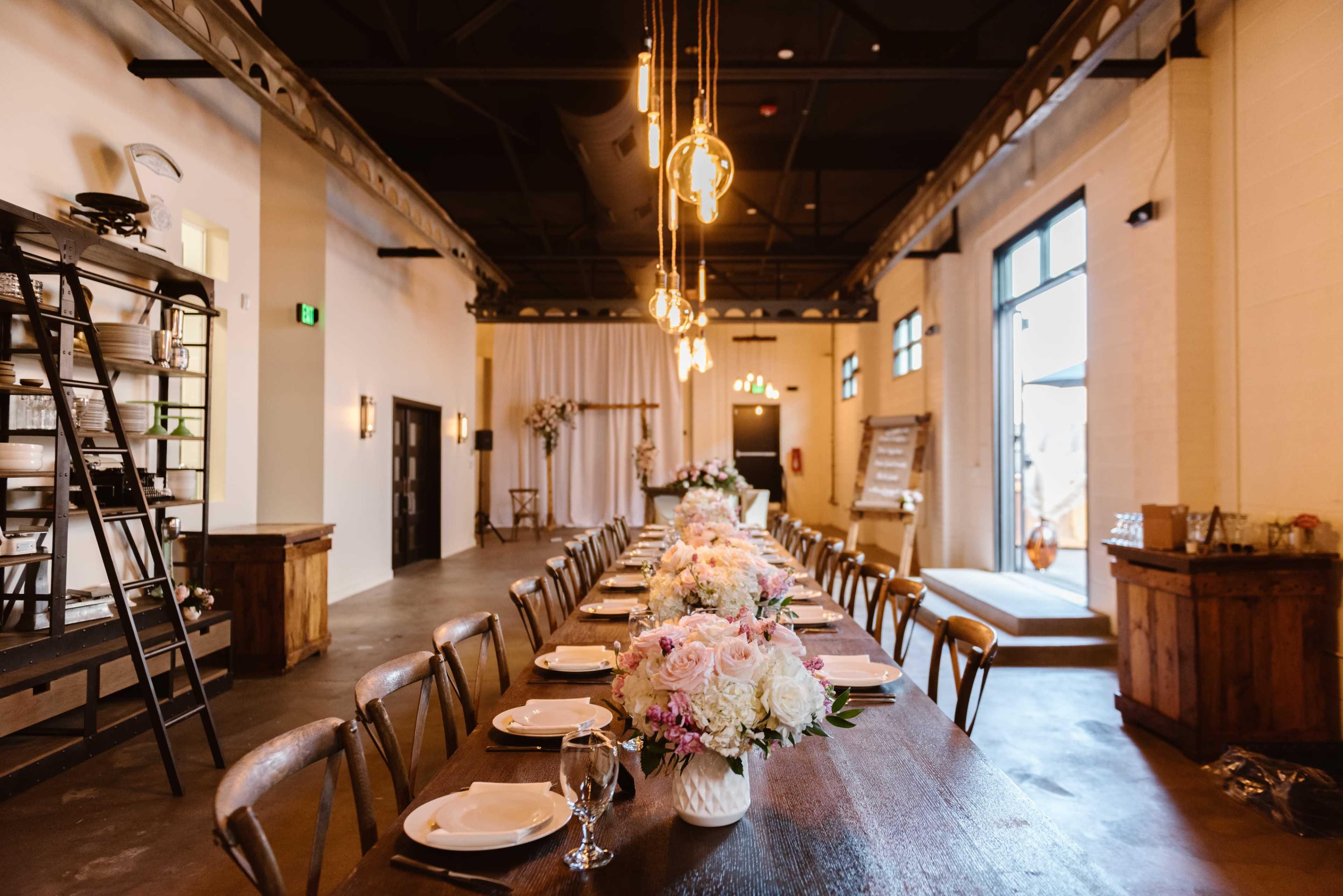 A long dining table is set with plates and floral arrangements in a spacious, well-lit venue.