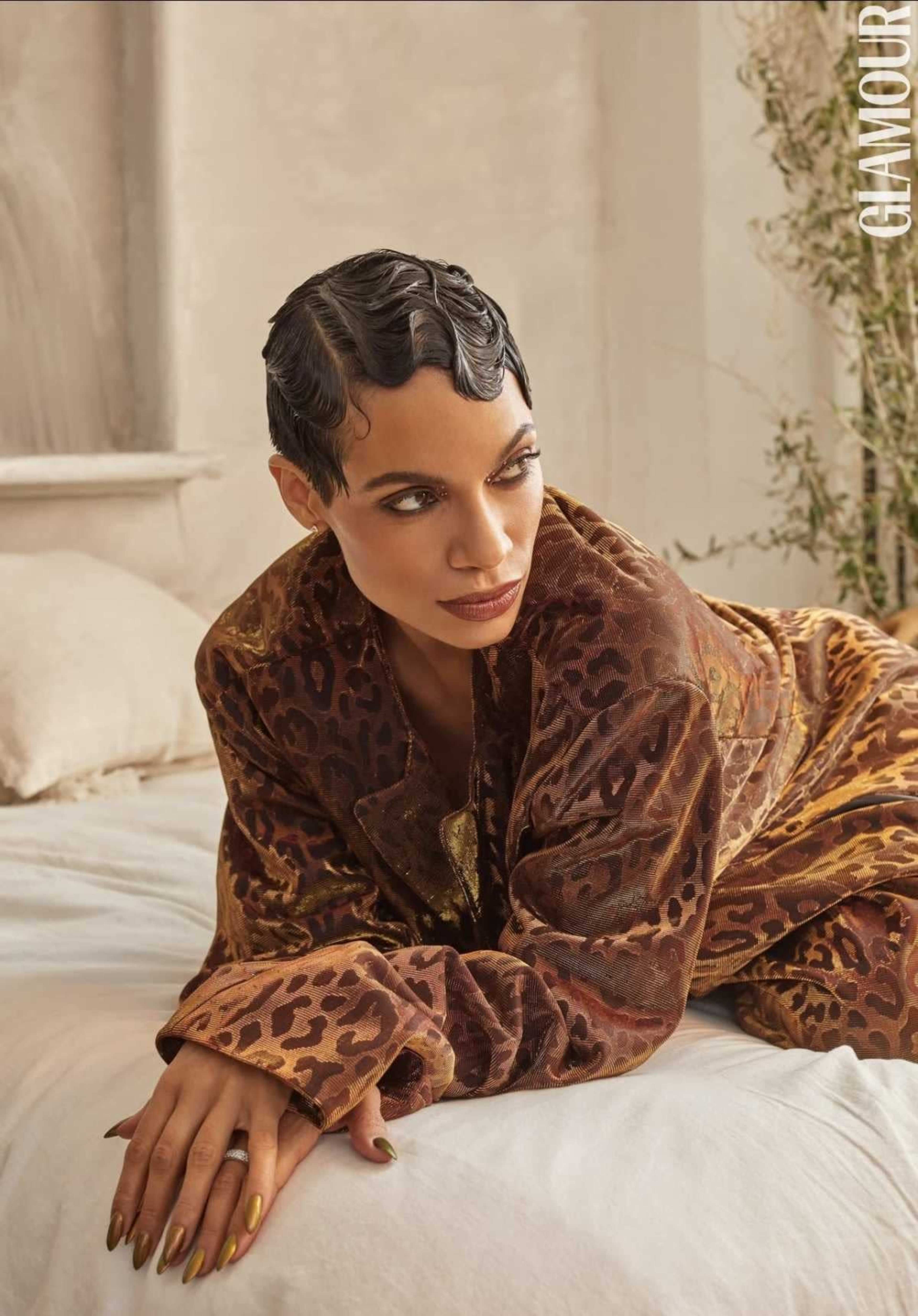 A person is reclining on a bed, wearing a patterned brown robe and styled hair, with a neutral-toned, softly lit background.