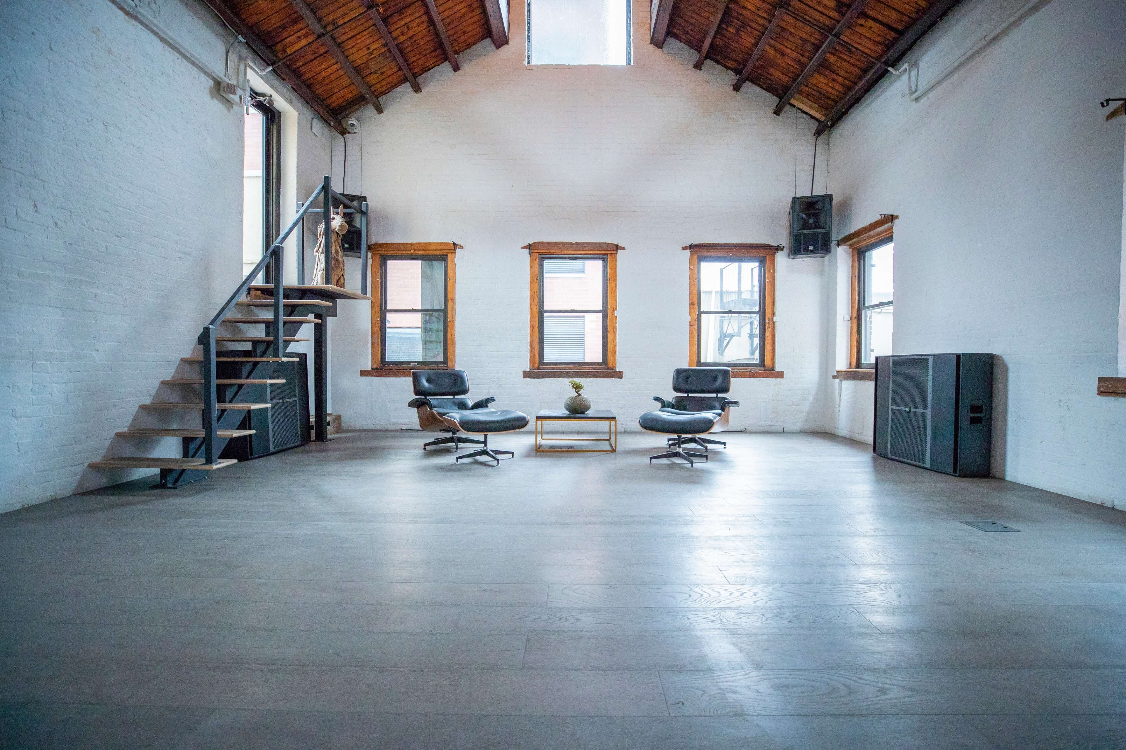 A spacious room with wooden beams, featuring two black chairs, a small table, and a staircase leading to a second level.