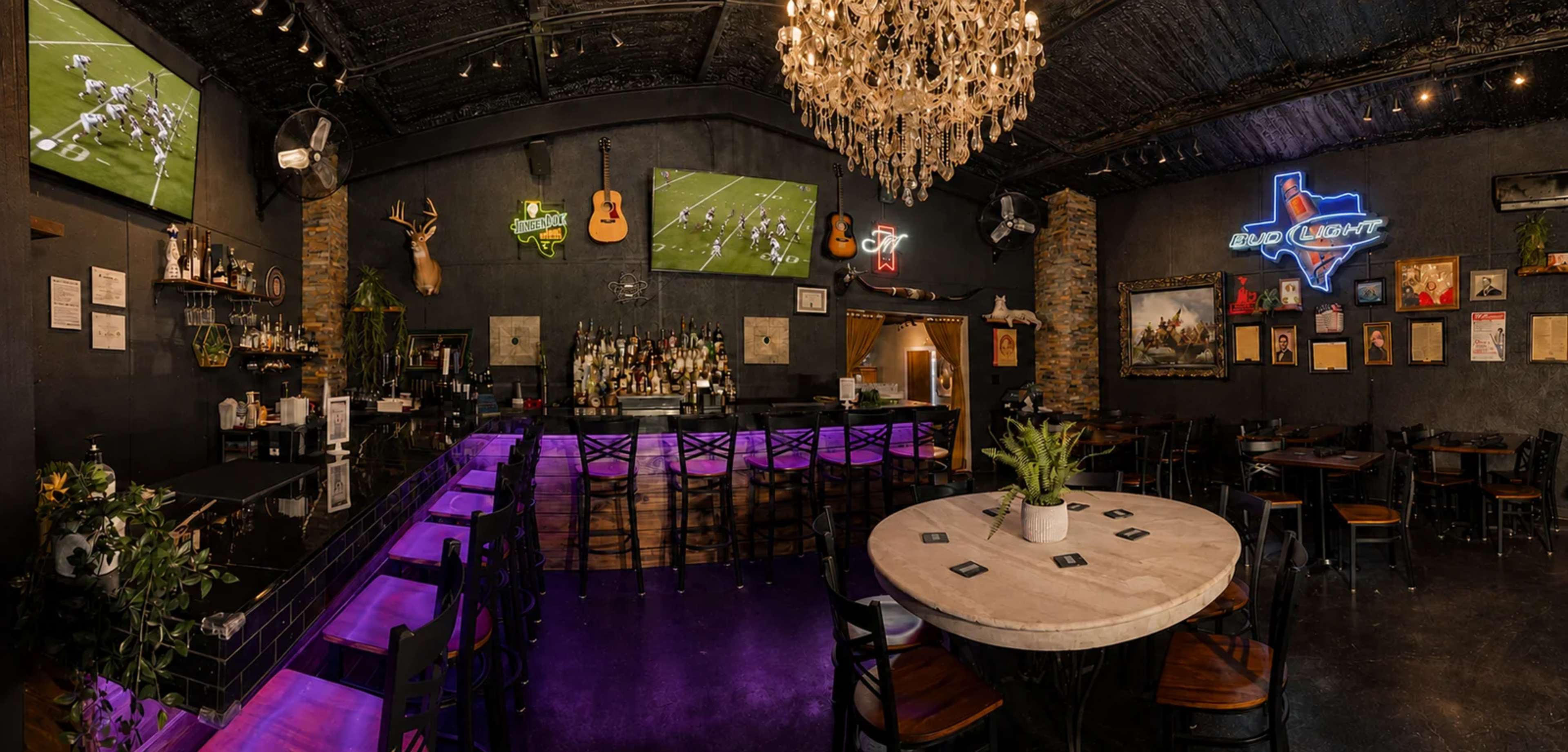 The image shows a well-decorated bar and dining area with a central round table, a long bar adorned with glowing purple lights, multiple televisions displaying a sports game, and various decorative items on the walls.