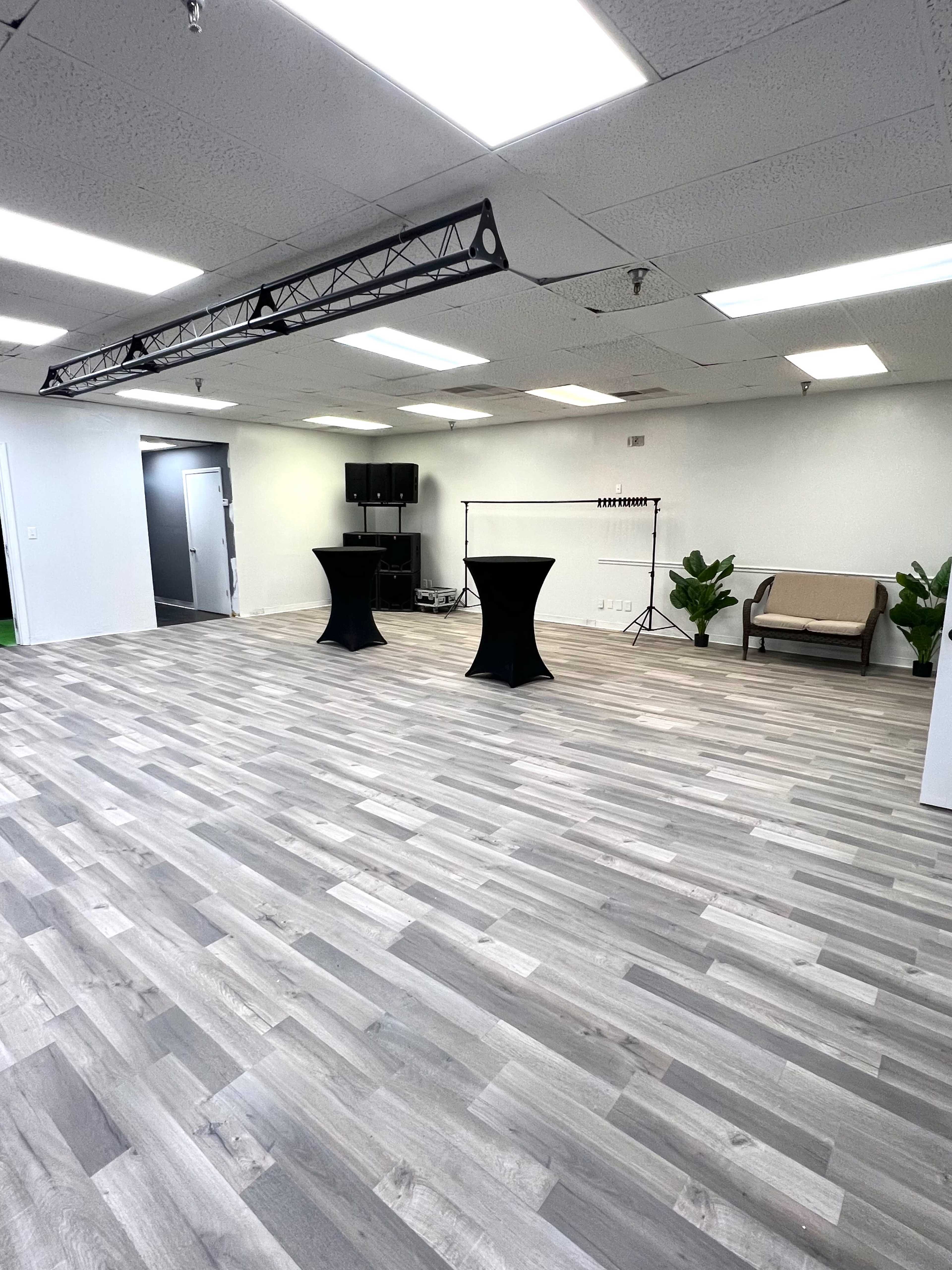 The spacious room features gray flooring, two black cocktail tables, a couch, and some greenery, with various lighting fixtures overhead.