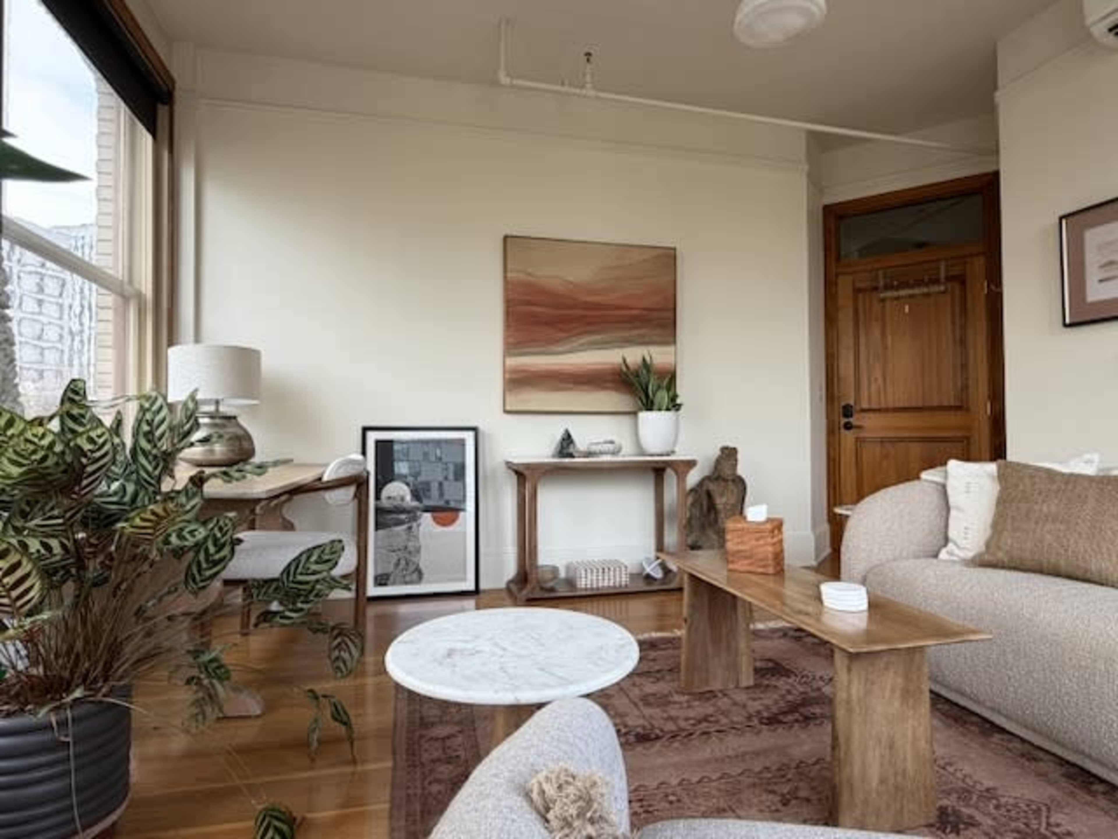 The image shows a cozy living room with a wooden door, a small marble coffee table, and decorative elements including plants and artwork.