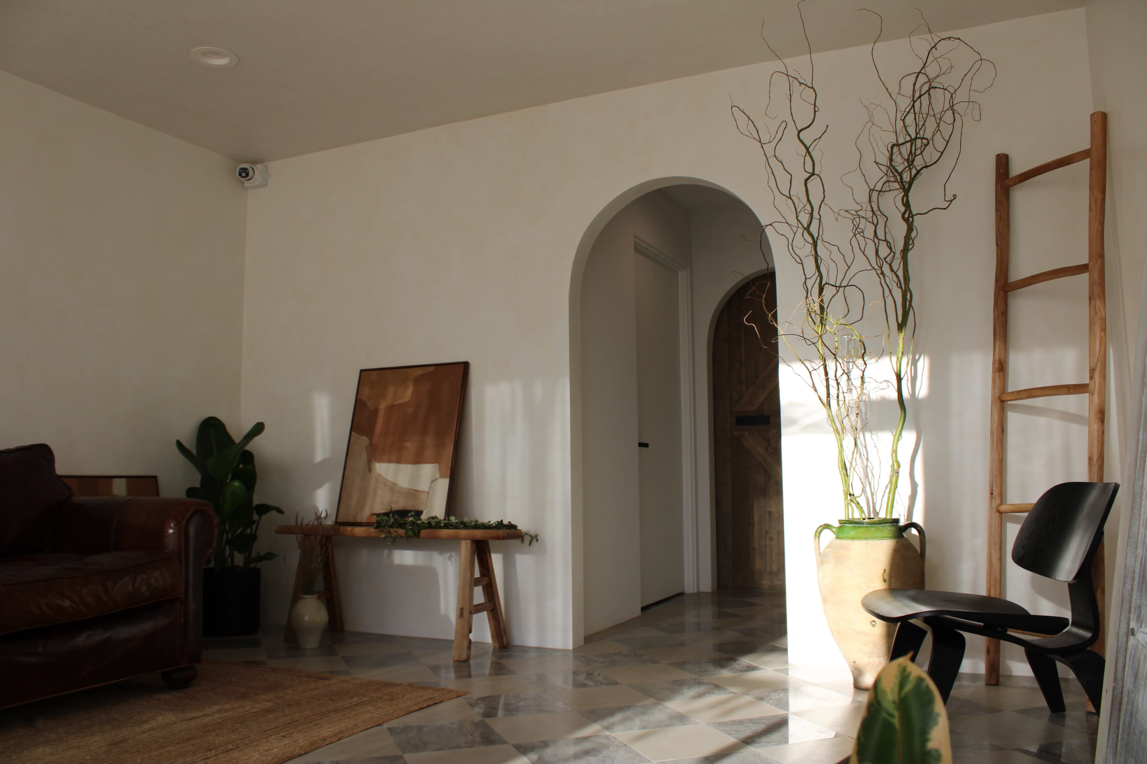 A sunlit interior features a leather sofa, a wooden table with decor, a tall plant, and a ladder against the wall, leading to an arched doorway.