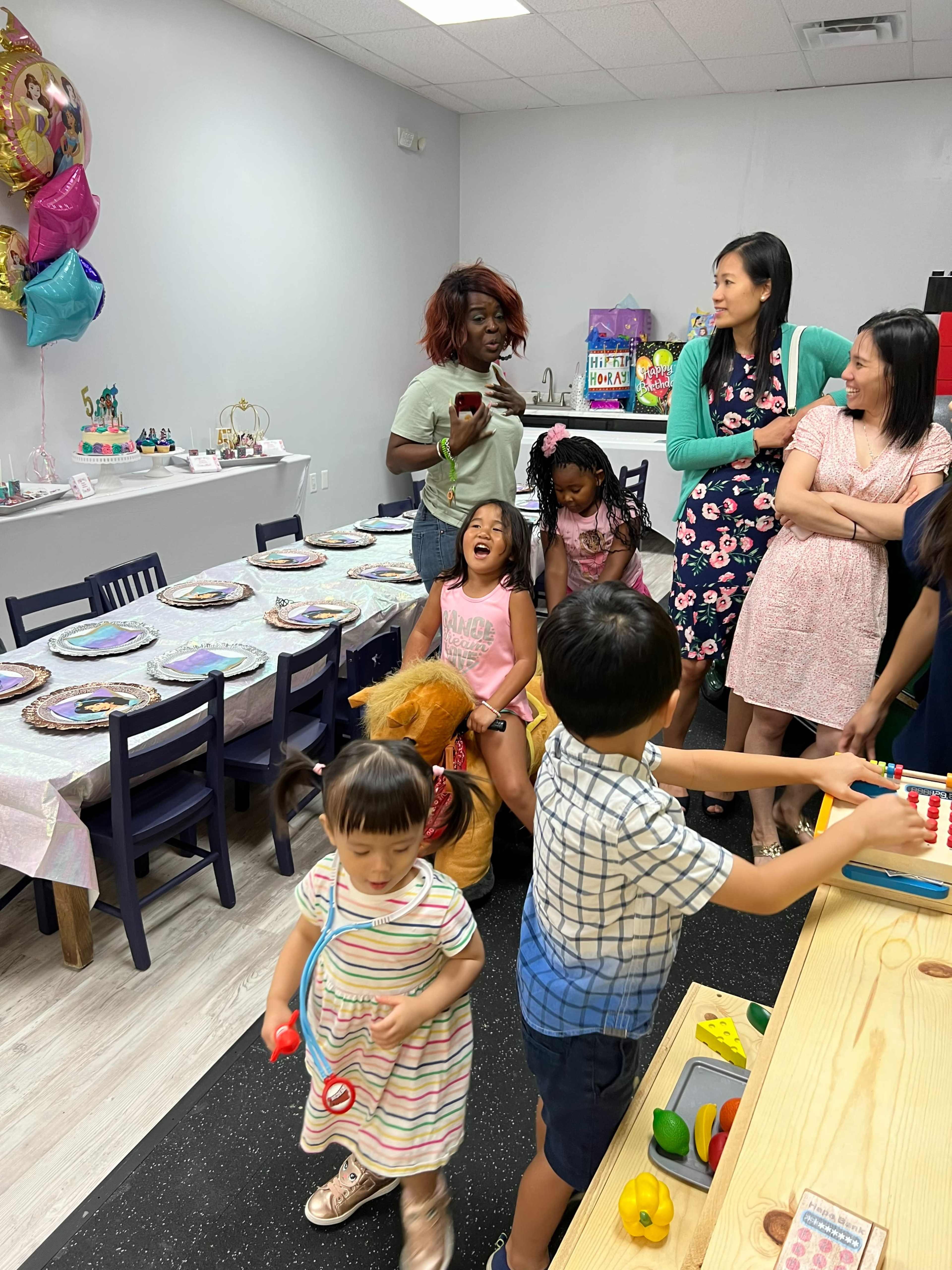 A group of children and adults are gathered in a decorated party space with tables set for a celebration.