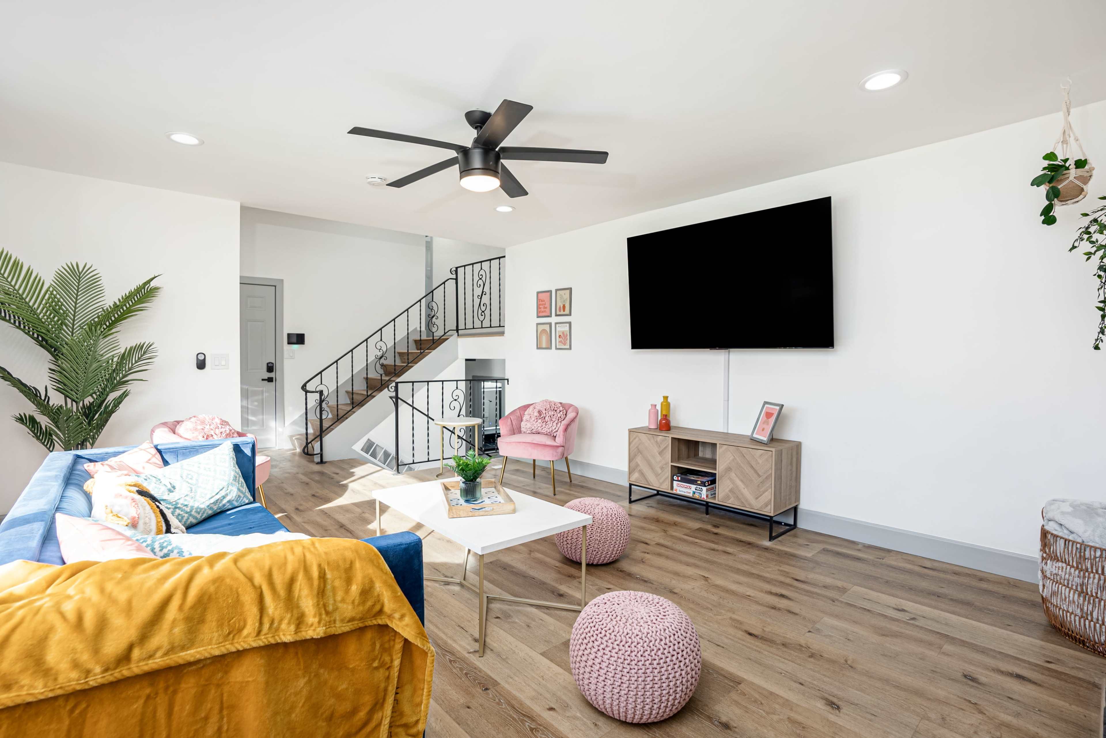 A modern living room featuring a blue sofa, pink armchairs, a wooden TV stand, and a staircase in the background.