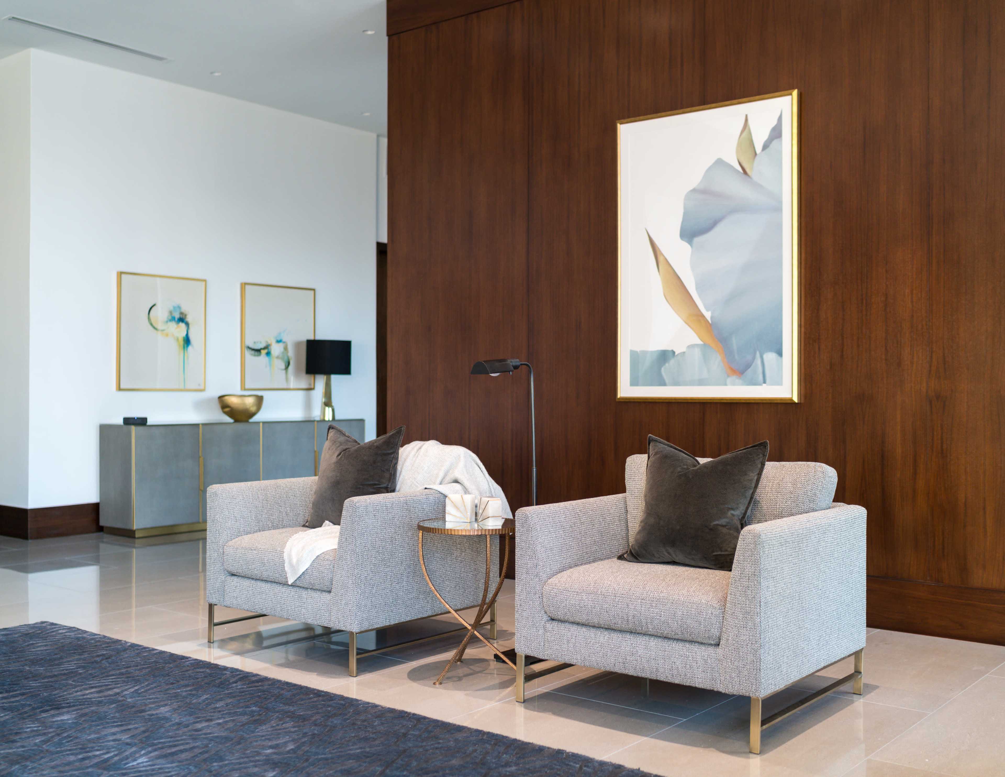 A pair of gray upholstered armchairs with decorative pillows is positioned next to a small side table in a modern lobby with a wooden wall and framed artwork.