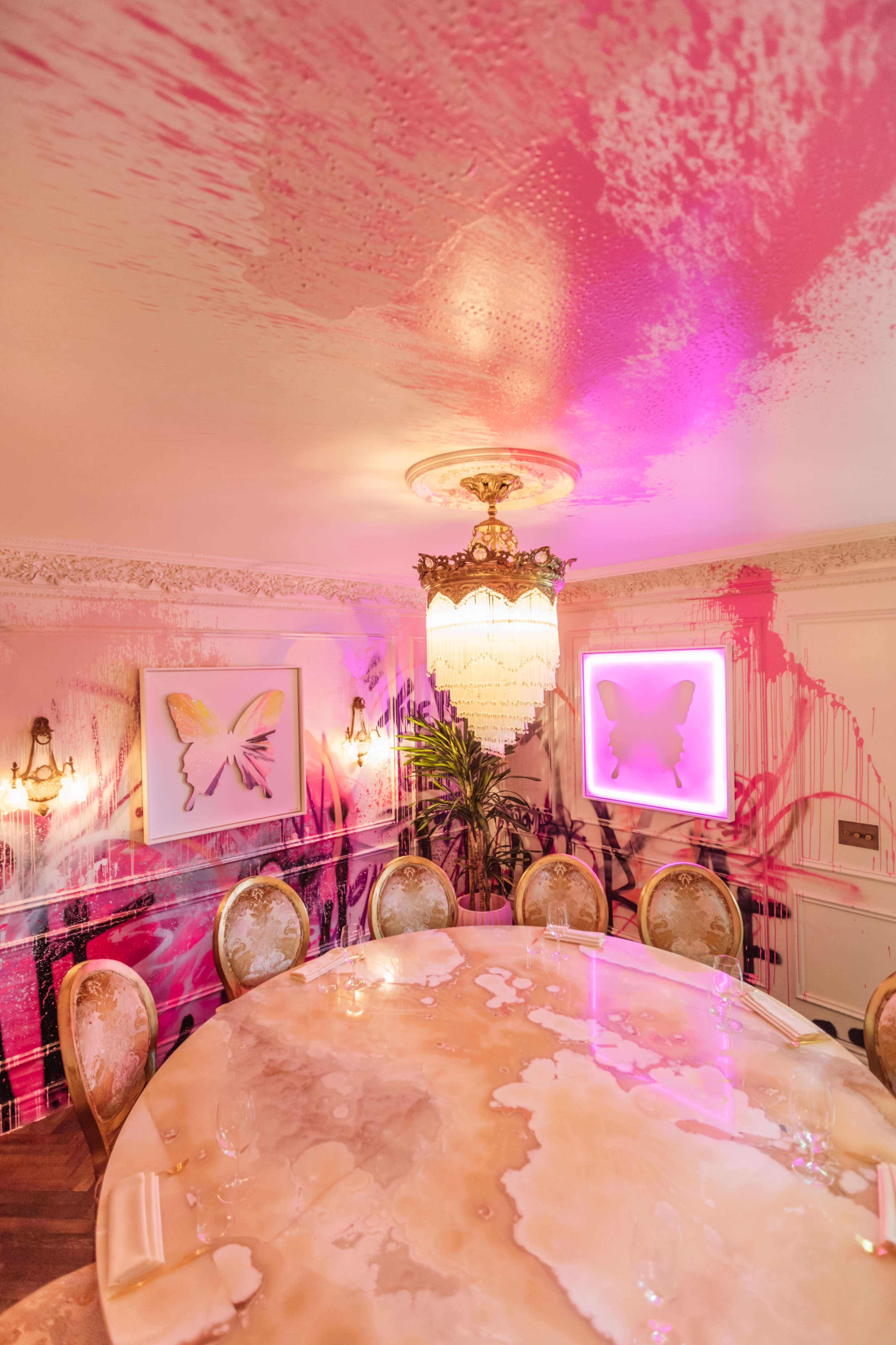 A dining room features a large round table surrounded by upholstered chairs, with butterfly artwork and vibrant wall decor highlighted by a chandelier.