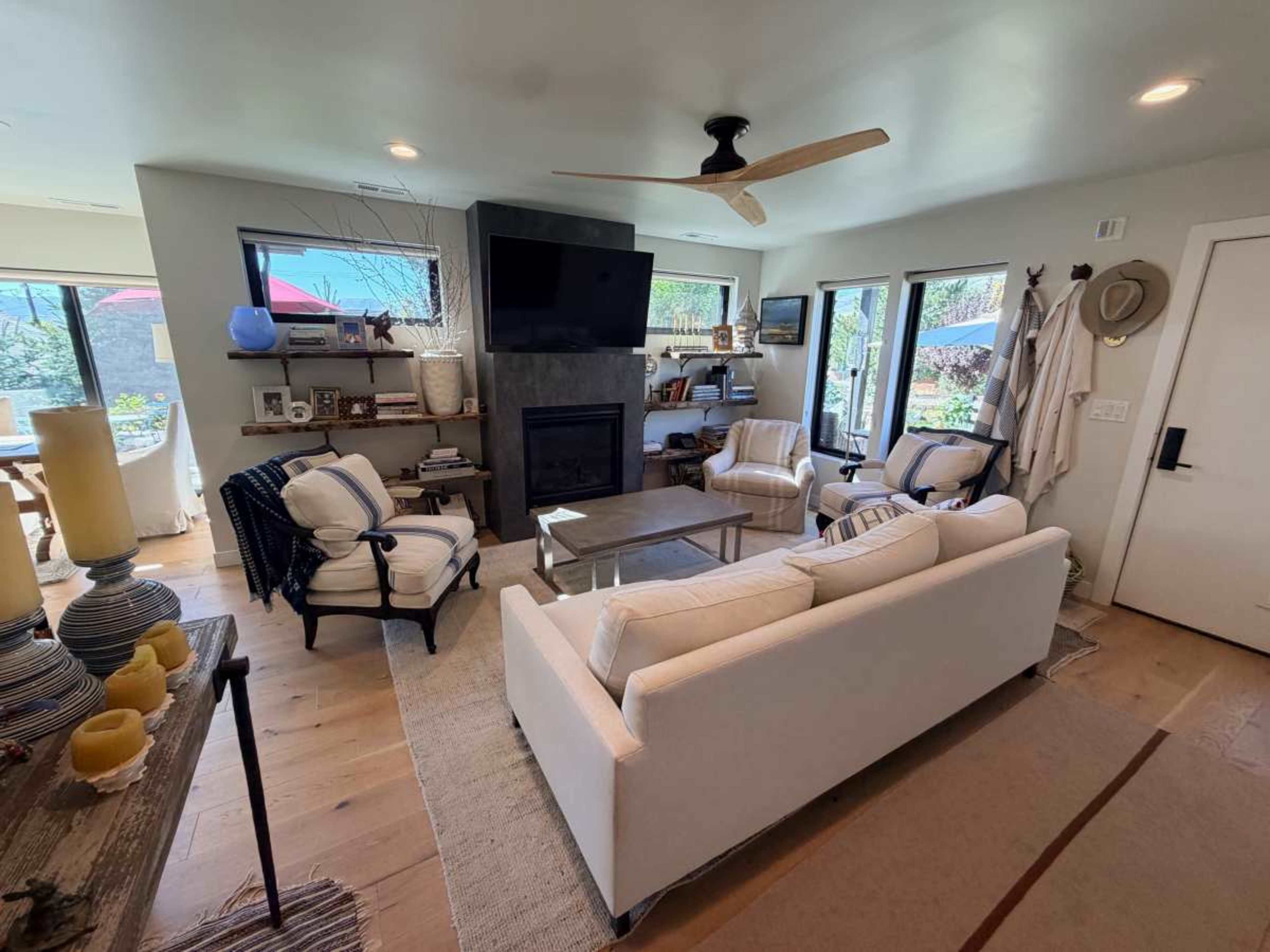 The image shows a modern living room featuring a light-colored sofa, armchairs, a coffee table, and a stone fireplace, with large windows providing natural light.