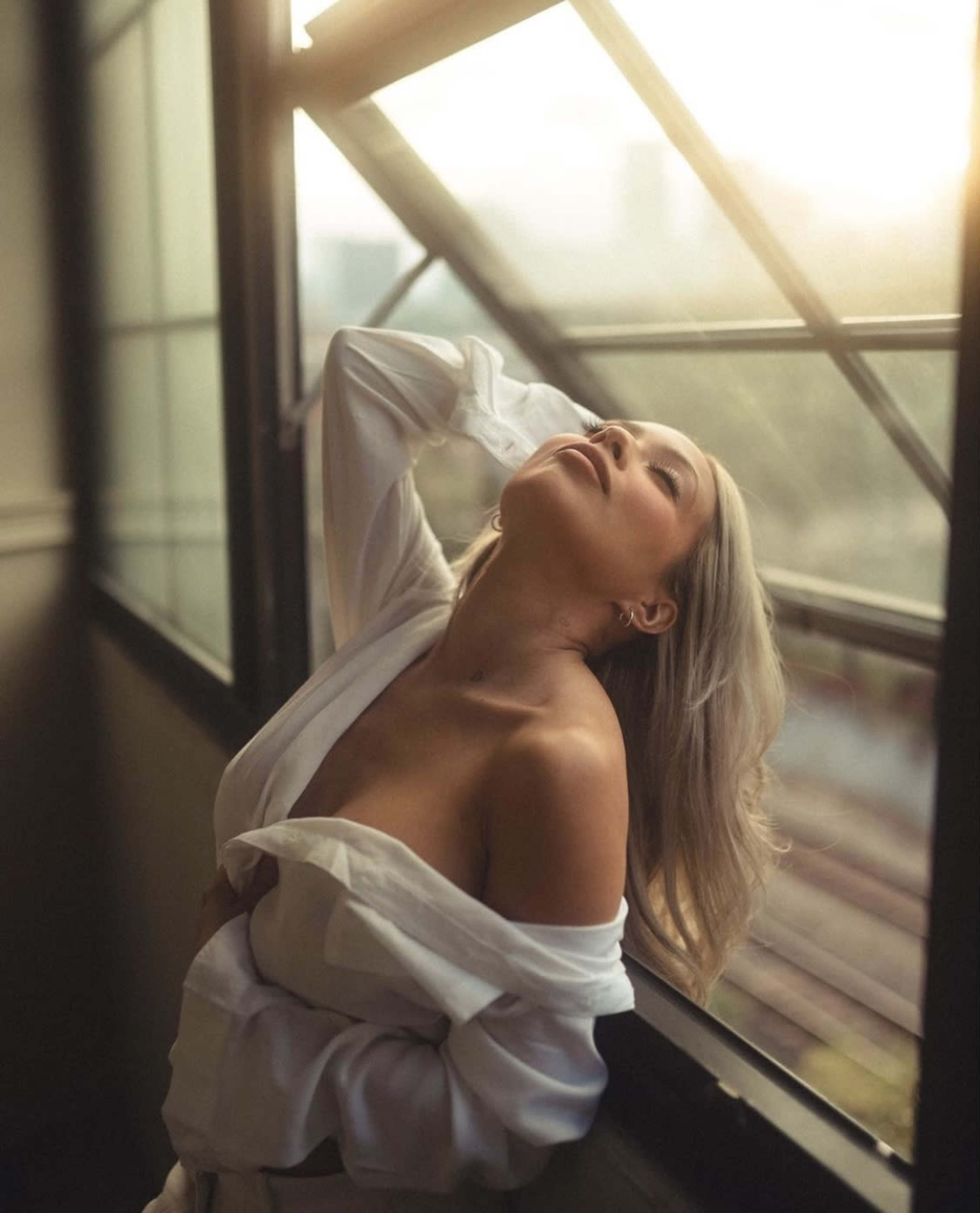 A woman in a white shirt leans against a large window, gazing upwards with sunlight streaming in.