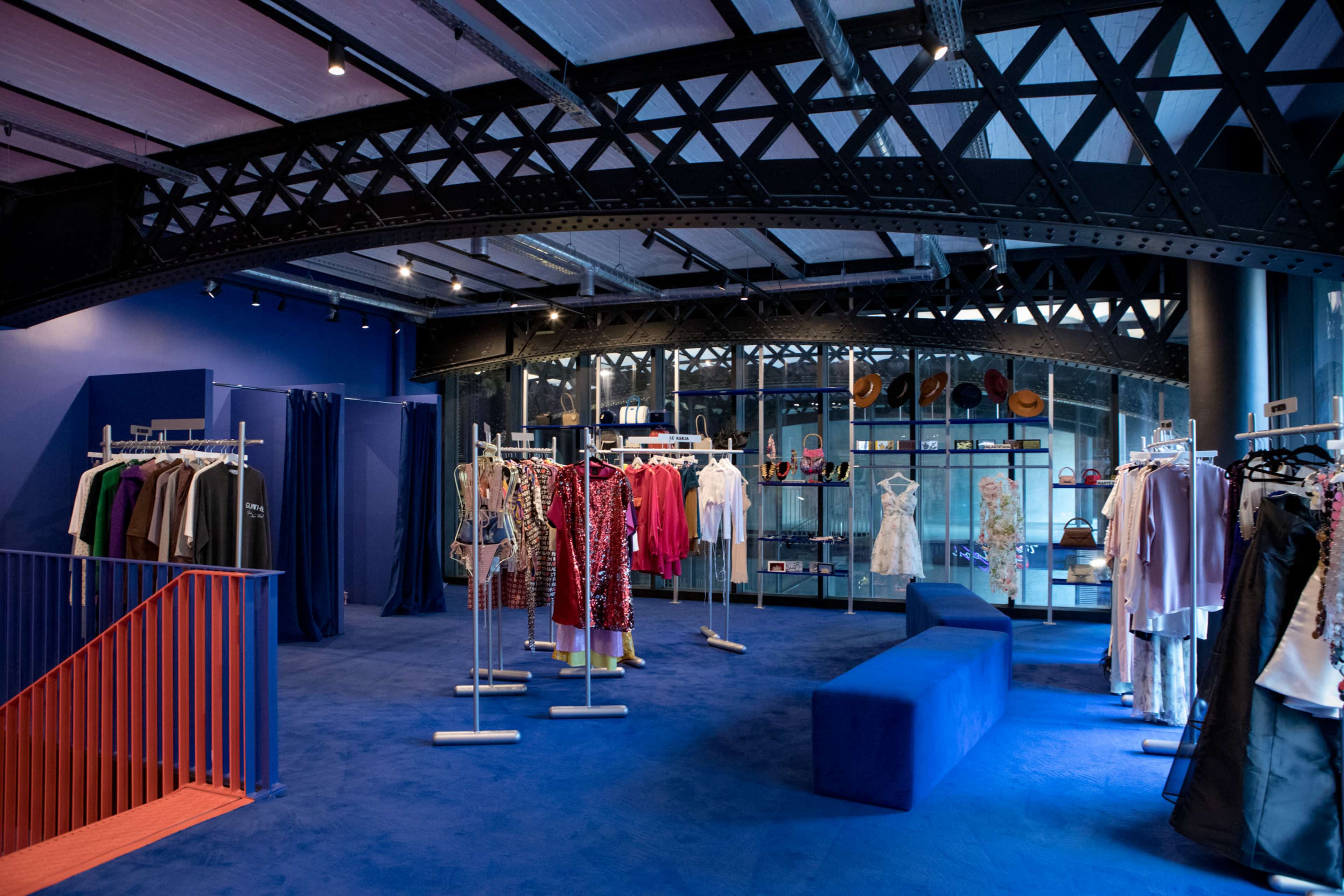 The image shows a modern clothing store interior with blue carpeting, clothing racks displaying a variety of garments, and large windows revealing a view of a bridge structure.