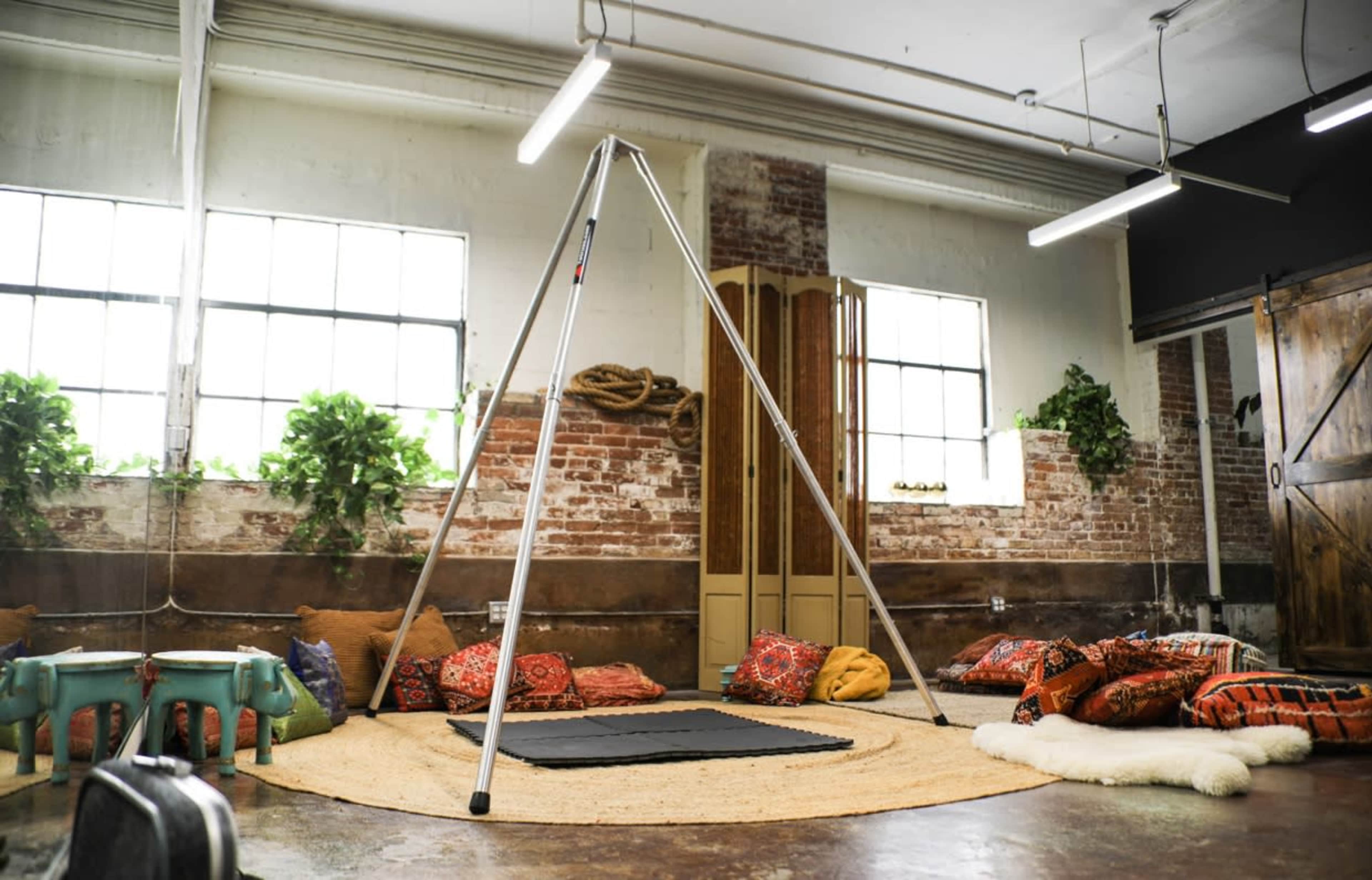 A spacious room with exposed brick walls, a geometric tripod structure, and various colorful floor cushions arranged around a circular rug.