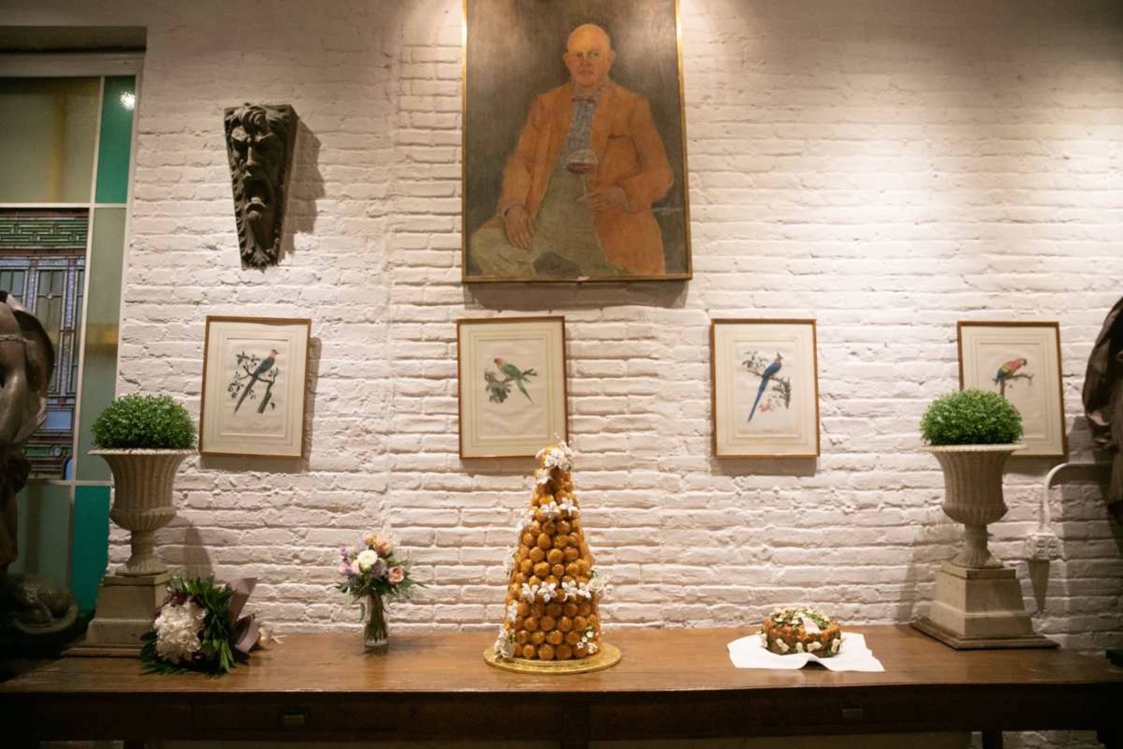 The image shows a decorative table displaying a tall croquembouche dessert, flanked by flower arrangements and artwork on a white brick wall.