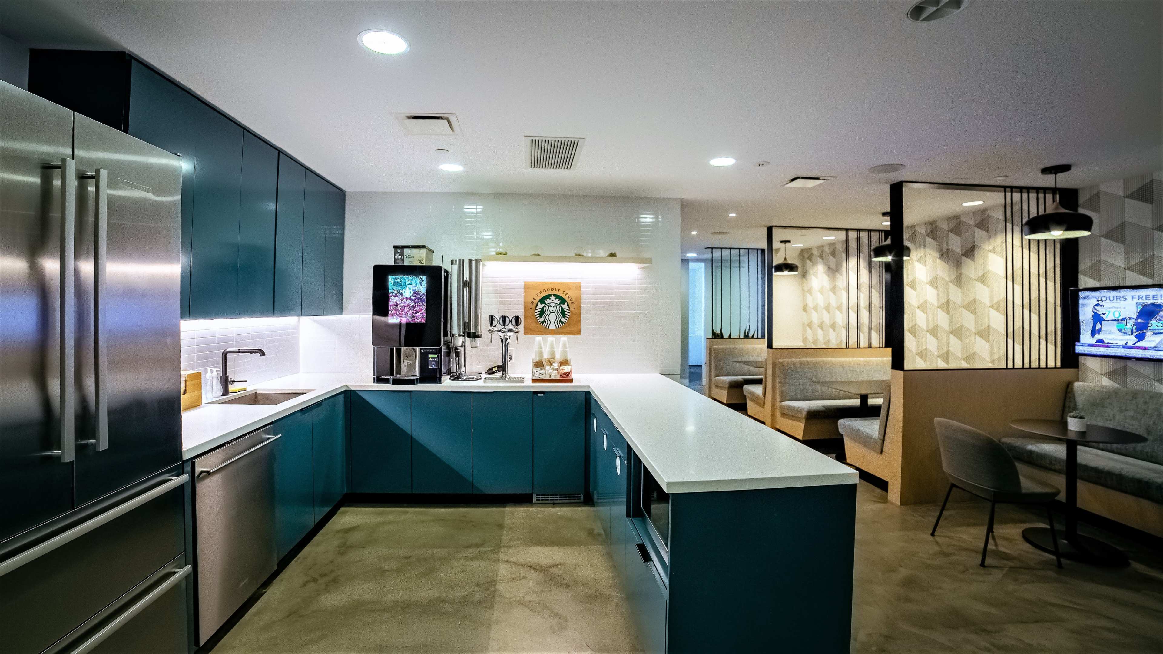 The image shows a modern kitchen area with teal cabinets, a coffee station, and a seating area featuring geometric walls and booth-style seating.