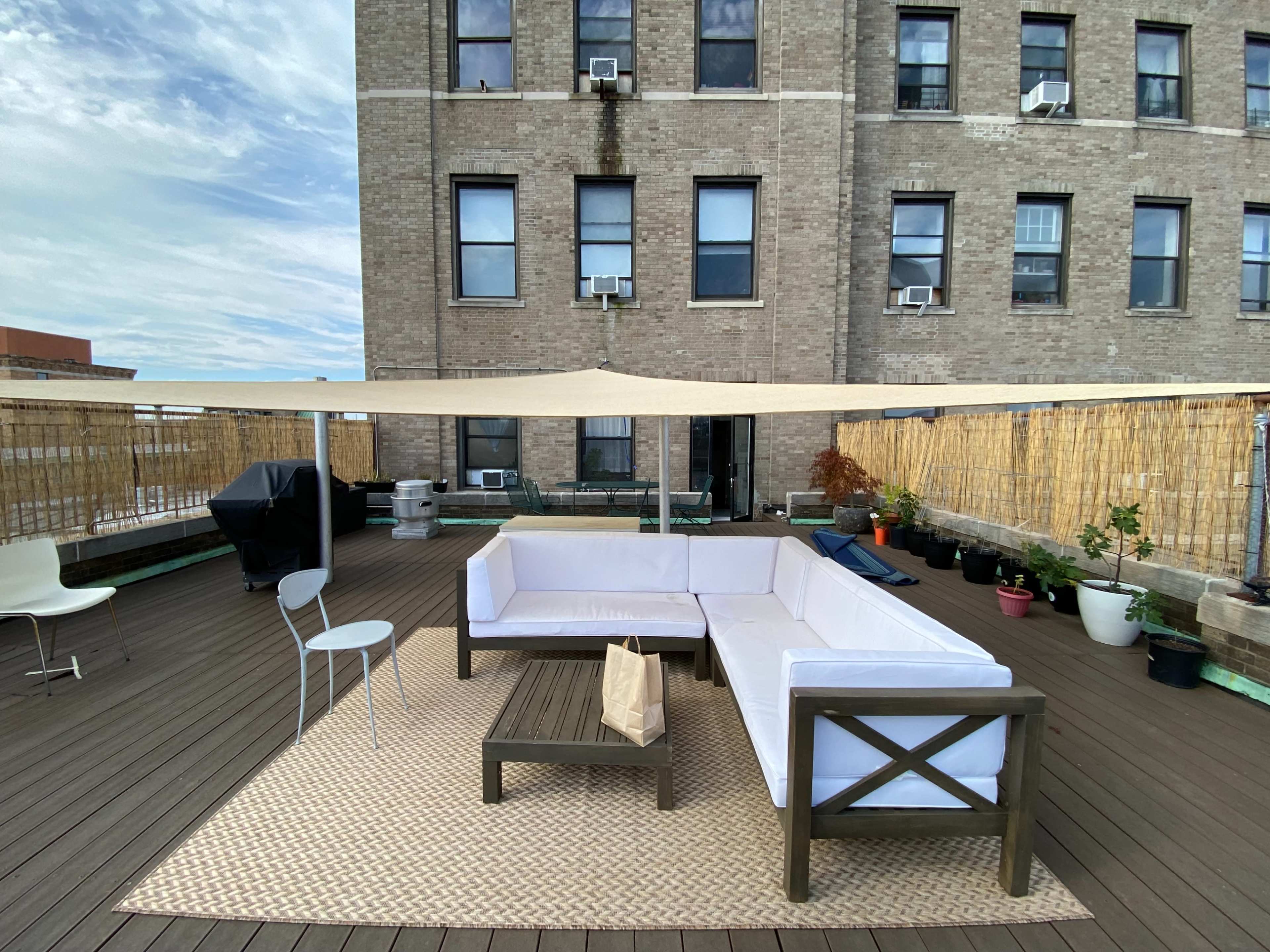 A rooftop patio features a sectional sofa, a coffee table, and potted plants under a canopy, with an apartment building in the background.
