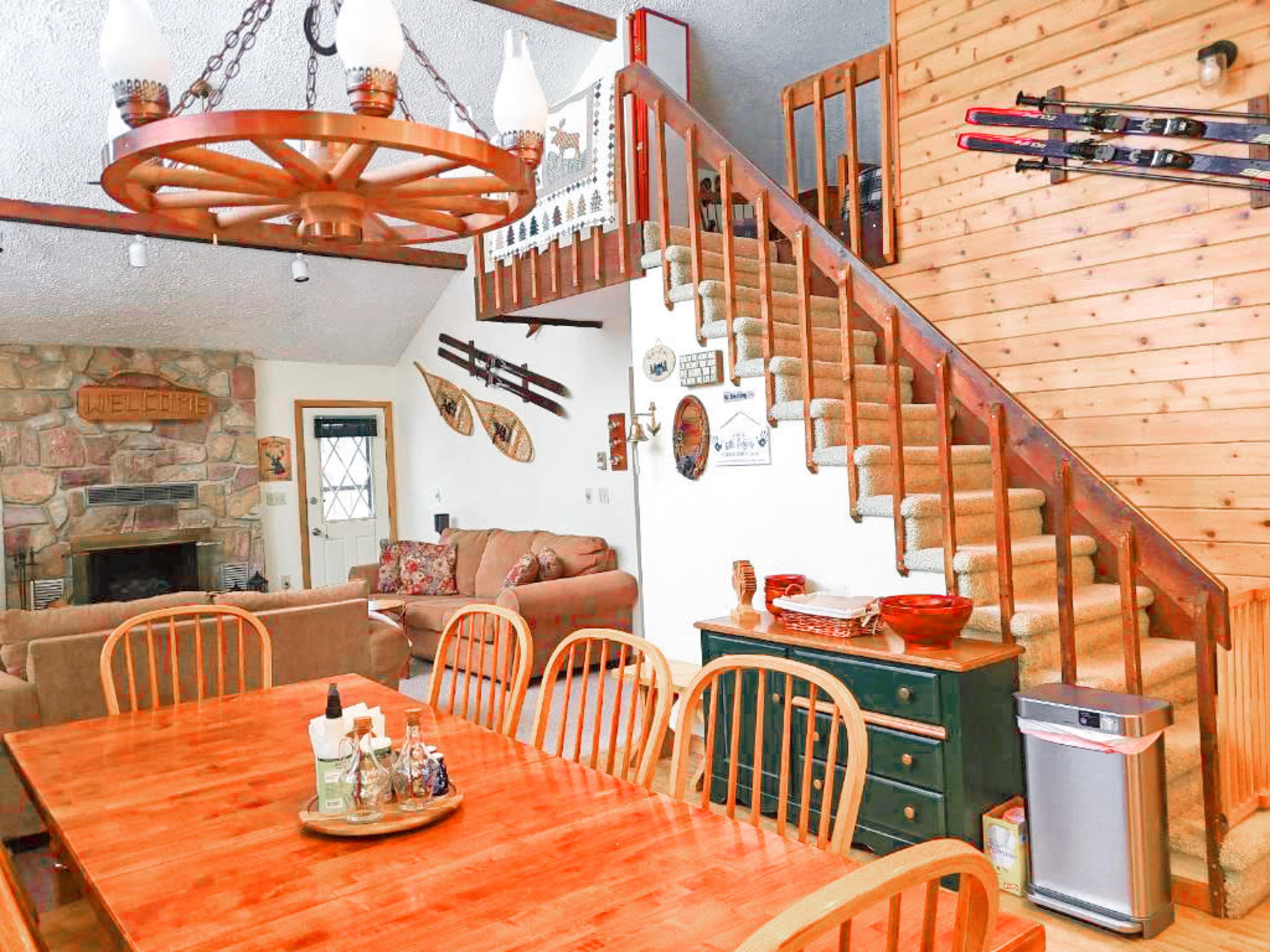 The image shows a rustic dining area with a wooden table, chairs, and a staircase leading to an upper level, adjacent to a living room with a stone fireplace.