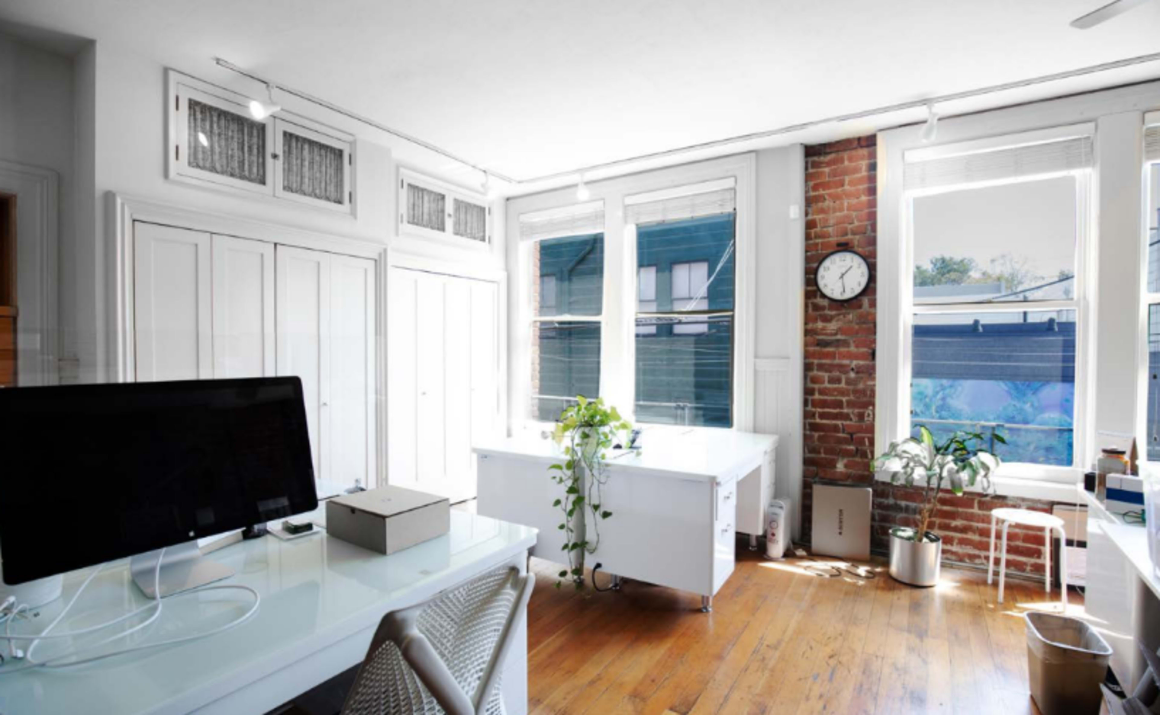 The image shows a well-lit office space featuring white furniture, exposed brick walls, and large windows.