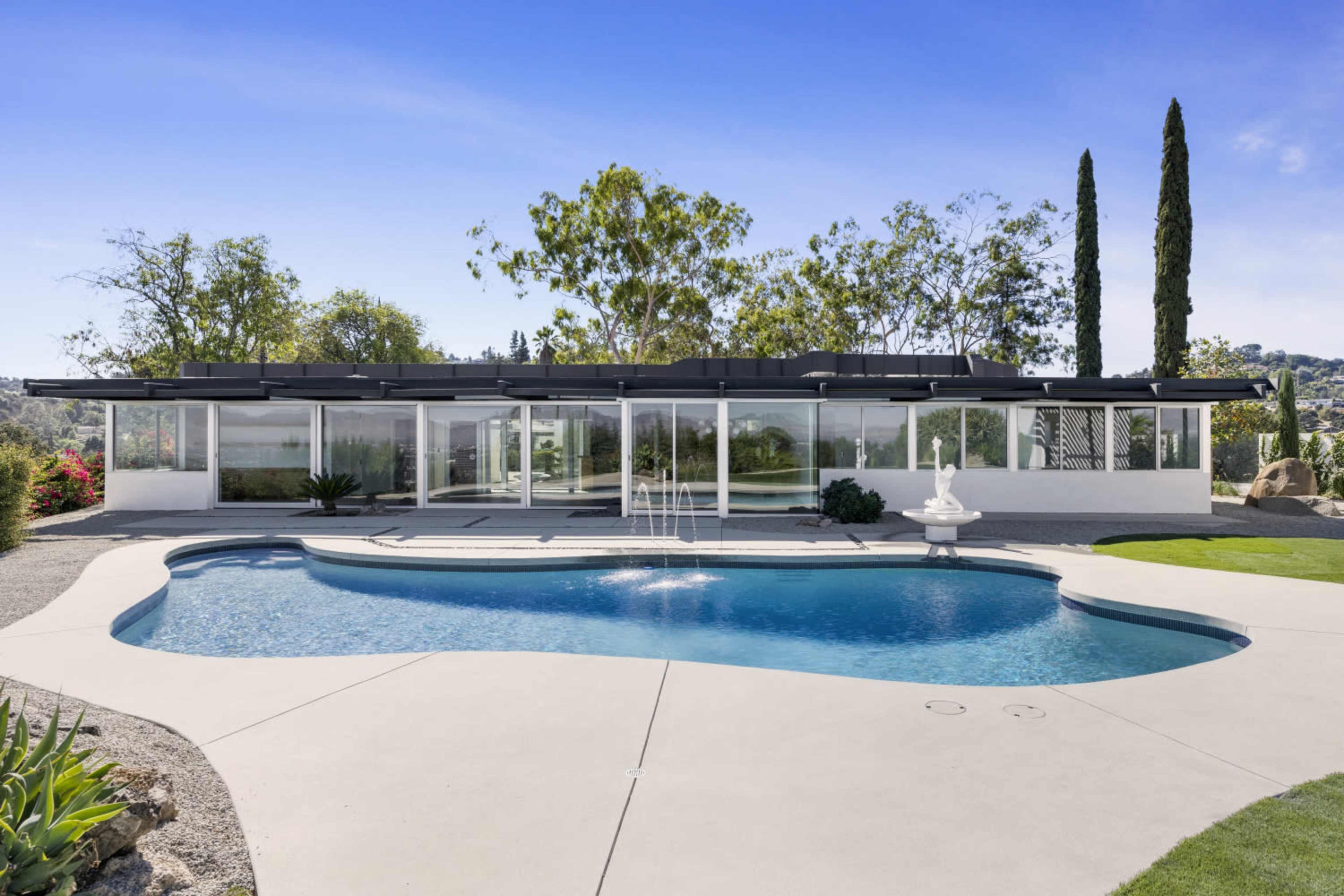The image shows a modern house with large glass windows surrounding a swimming pool, set against a backdrop of trees and landscaping.