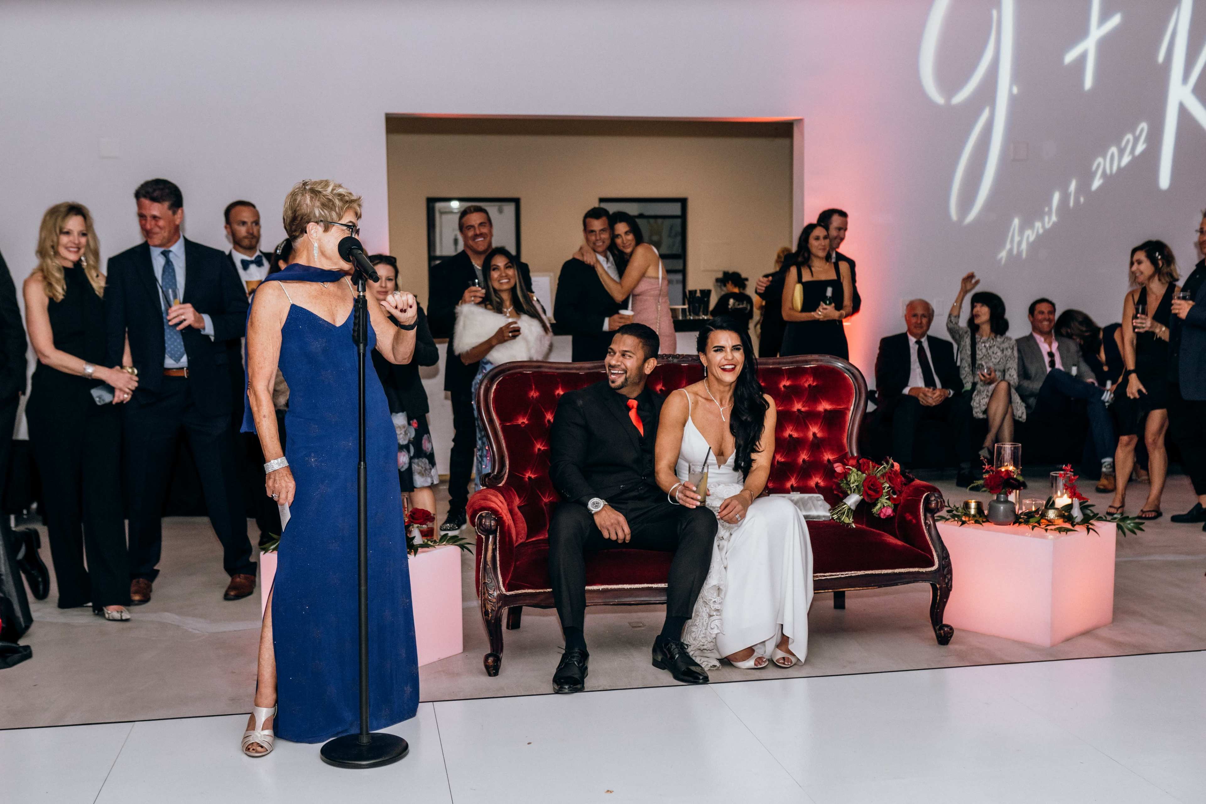 A woman speaks at a microphone while a couple sits on a red velvet couch in front of a crowd during a wedding reception.