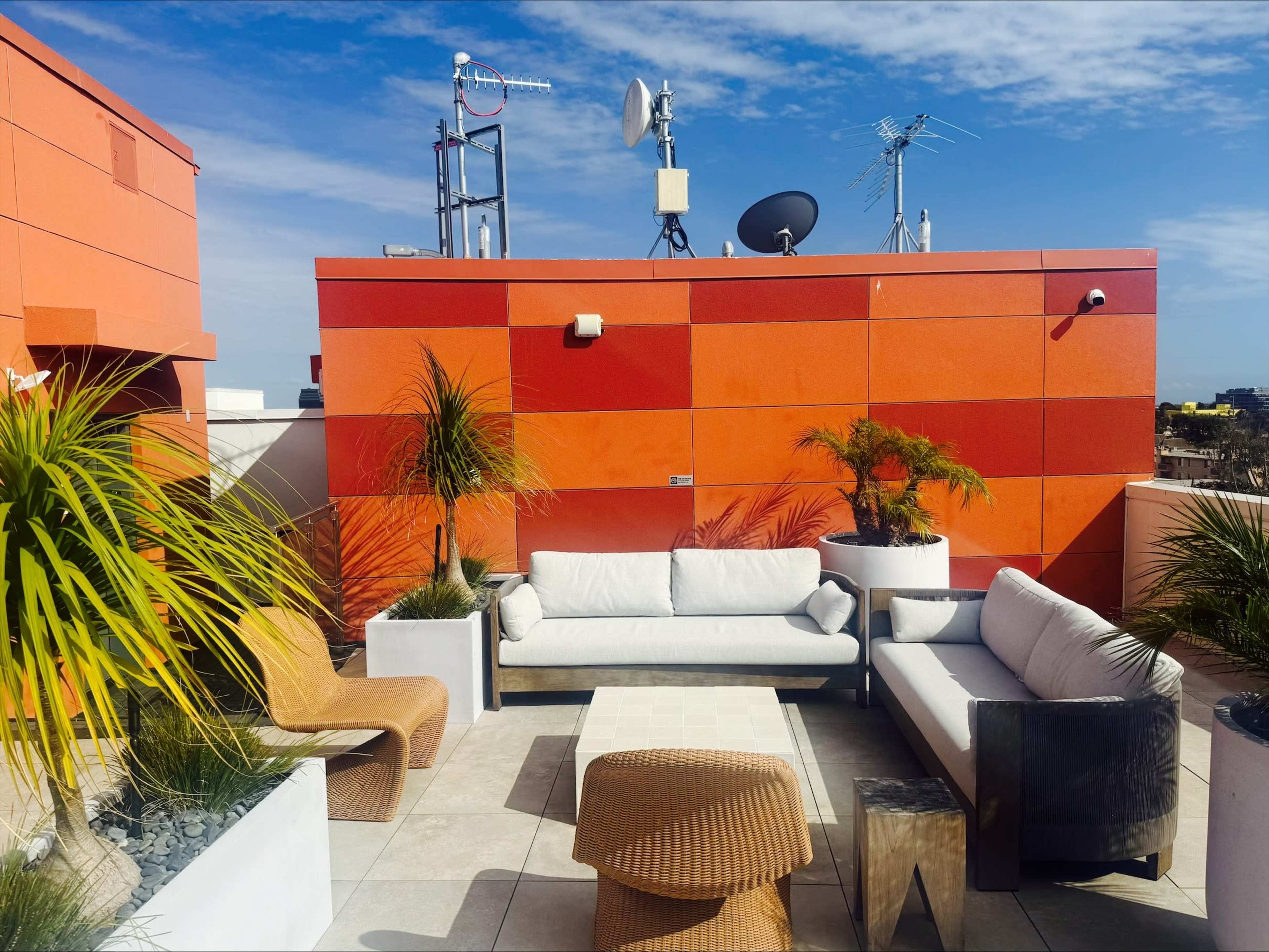 The image shows a rooftop lounge area featuring modern outdoor furniture, potted plants, and various antennas on adjacent buildings under a clear blue sky.