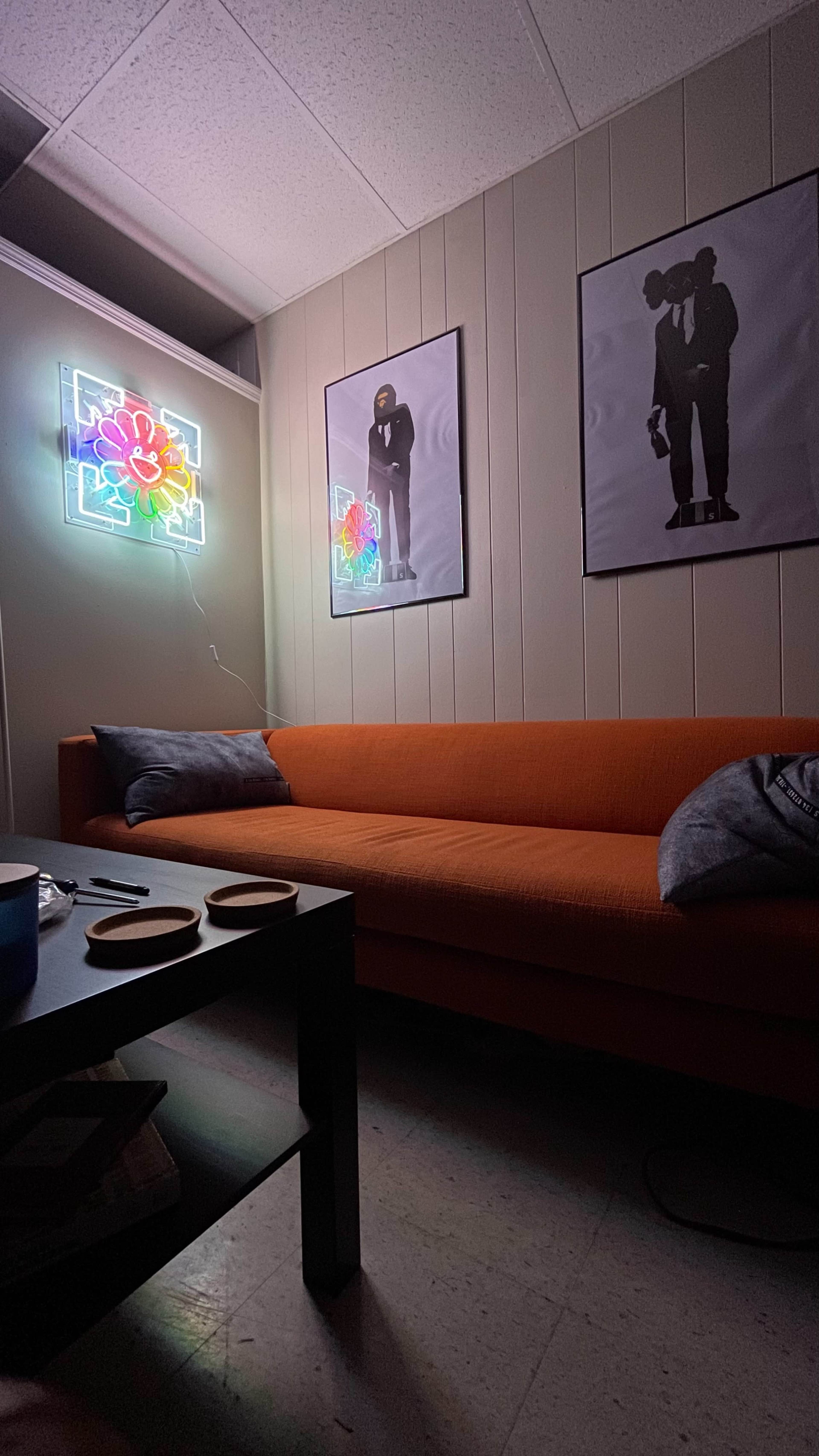 The image shows a dimly lit room featuring an orange couch, a black coffee table with coasters, and two framed posters on the walls, along with a neon floral light.