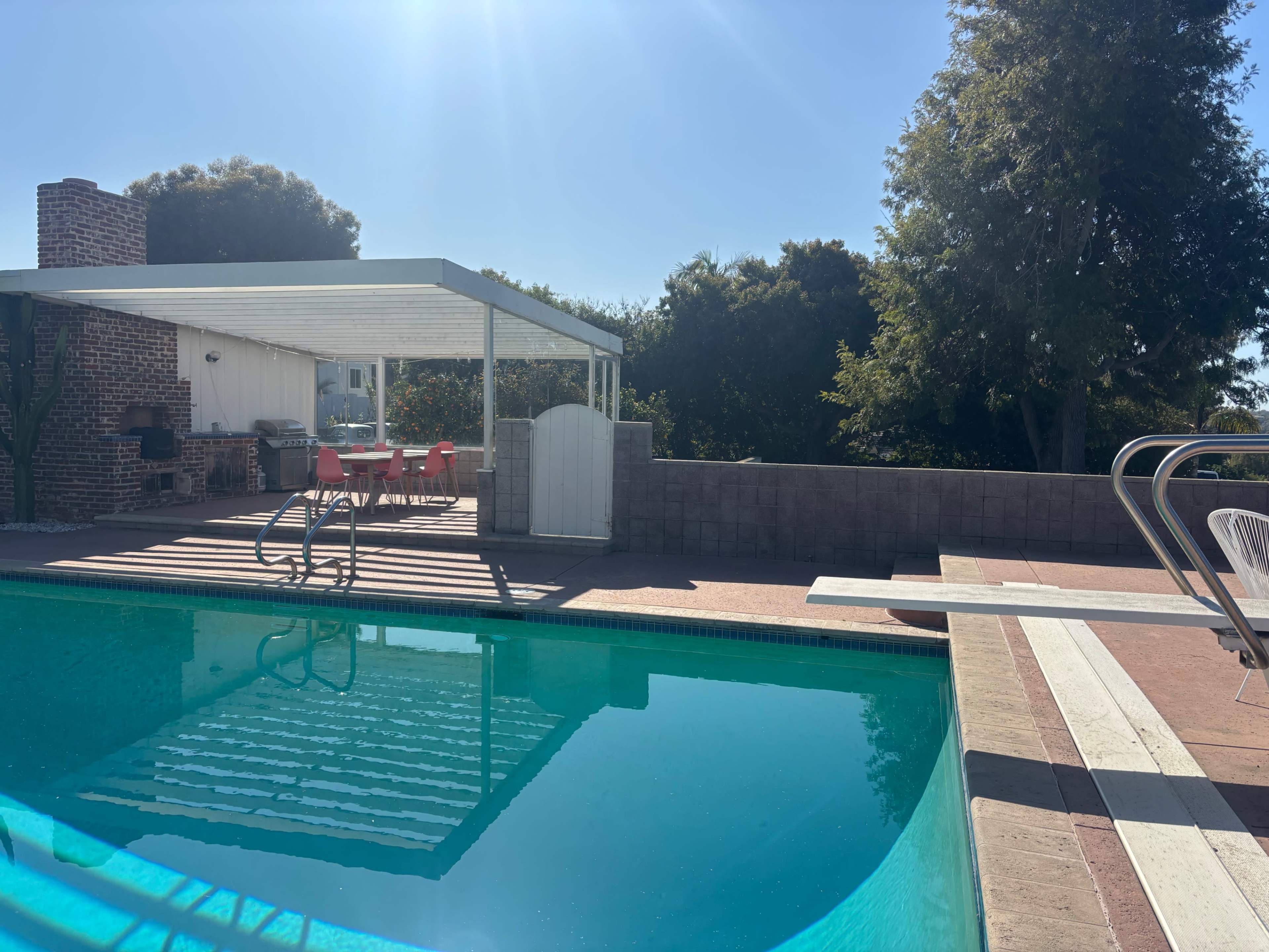 A swimming pool with a diving board is situated next to a house with a covered patio and a set of red chairs.